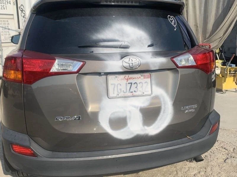 Back of a gray Toyota RAV4 with a California license plate, a white spray-painted peace symbol and ring on the trunk, and a small peace sign sticker on the rear window. The vehicle appears dirty and is parked outdoors near industrial surroundings.