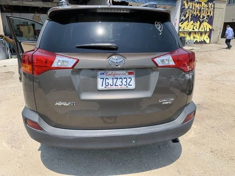 The image shows the back of a gray Toyota RAV4 Limited Auto vehicle with a California license plate reading 7GJZ332 parked on a dirt surface. There is a reflection of the sky on the rear window, and some buildings and graffiti are visible in the back