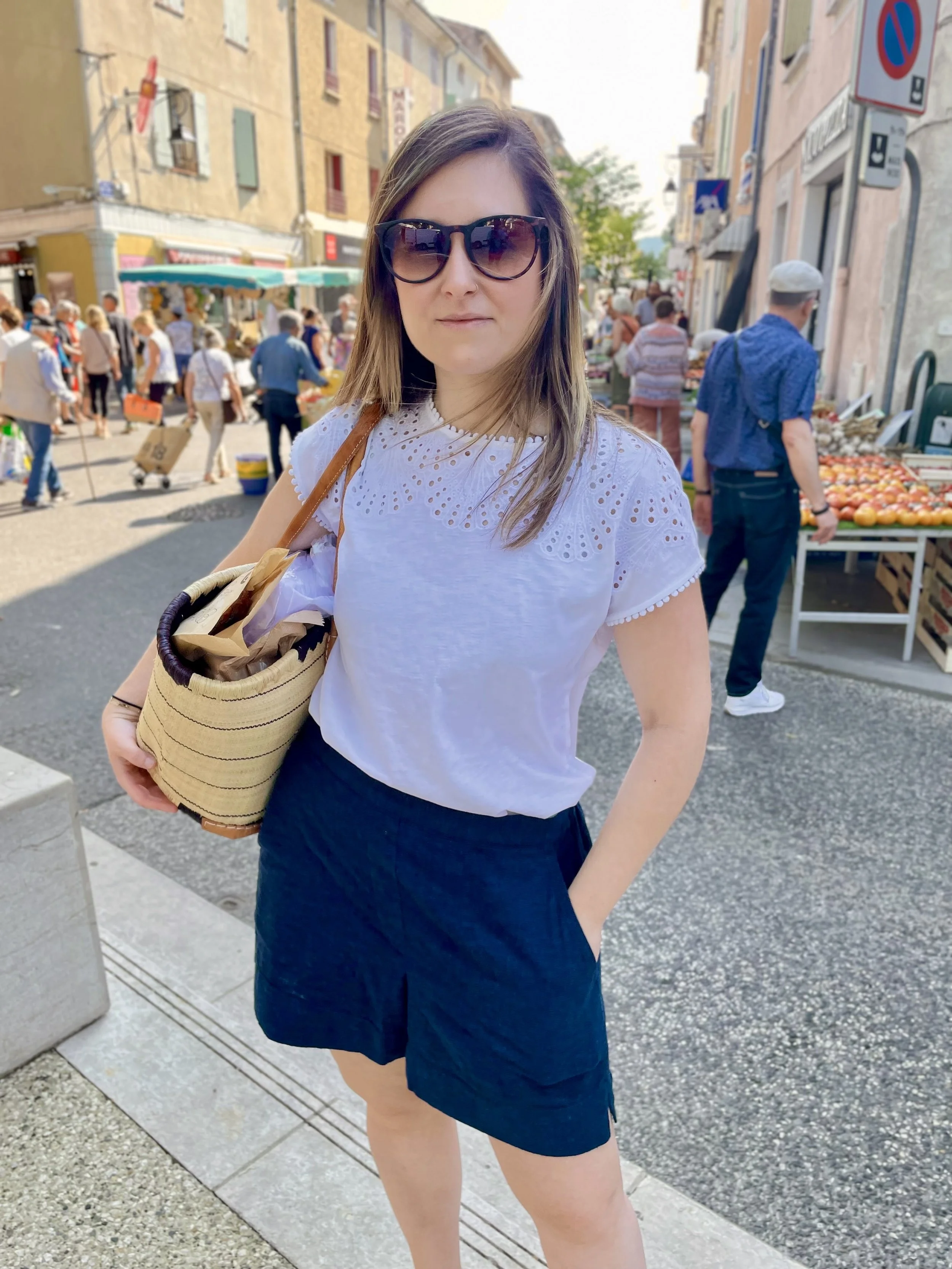 Woman with brown hair wearing sunglasses, a white eyelet top, and a navy skirt standing on a busy city street with market stalls and other people in the background.