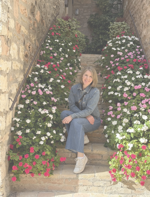 Woman in denim jacket and jeans sitting on brick stairs surrounded by pink, white, and purple flowers in planters.
