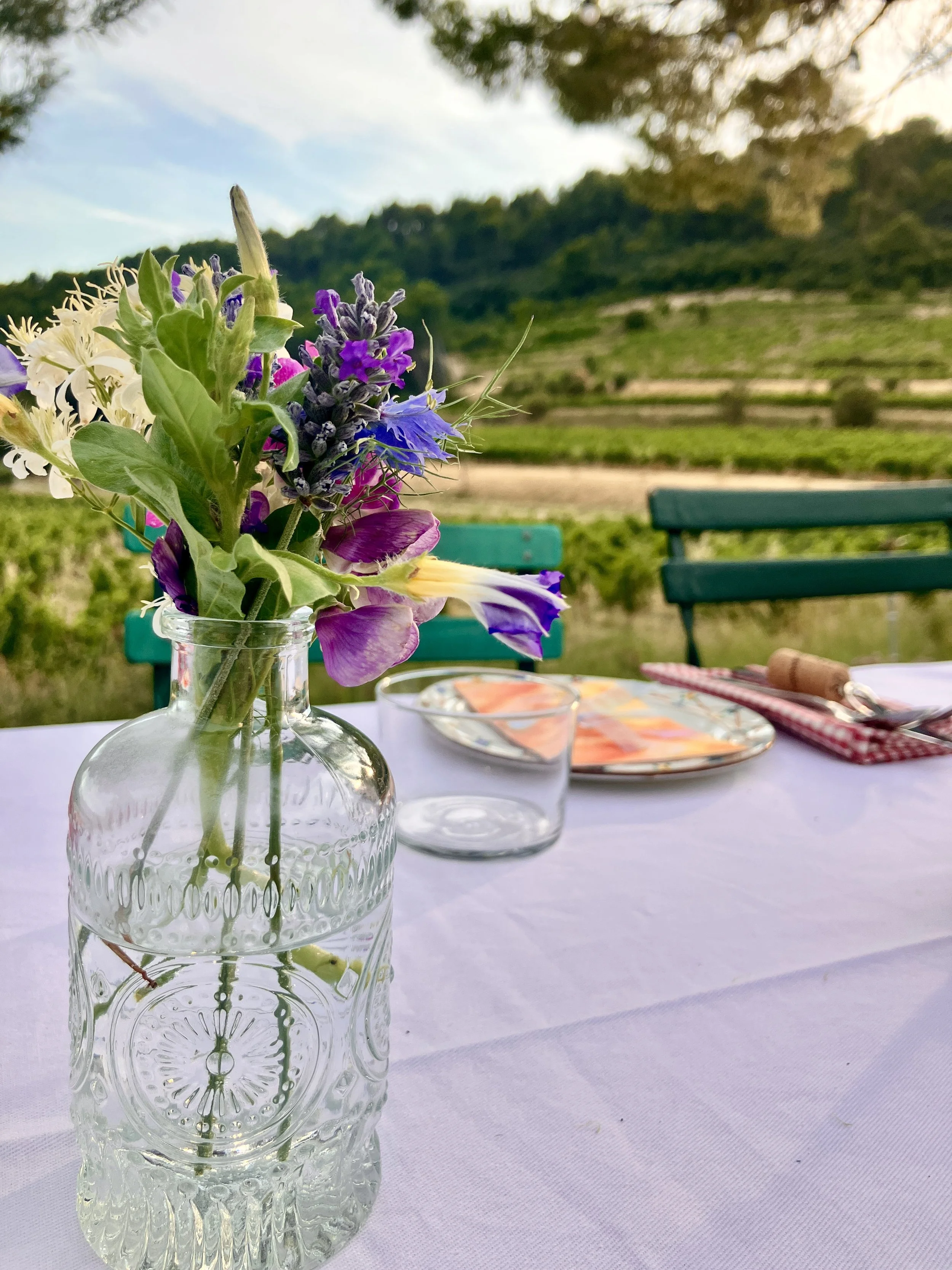 A clear glass vase with intricate patterns holds a bouquet of purple, pink, white, and green flowers on a white tablecloth, with a scenic outdoor background of green hills, trees, and a partly cloudy sky.