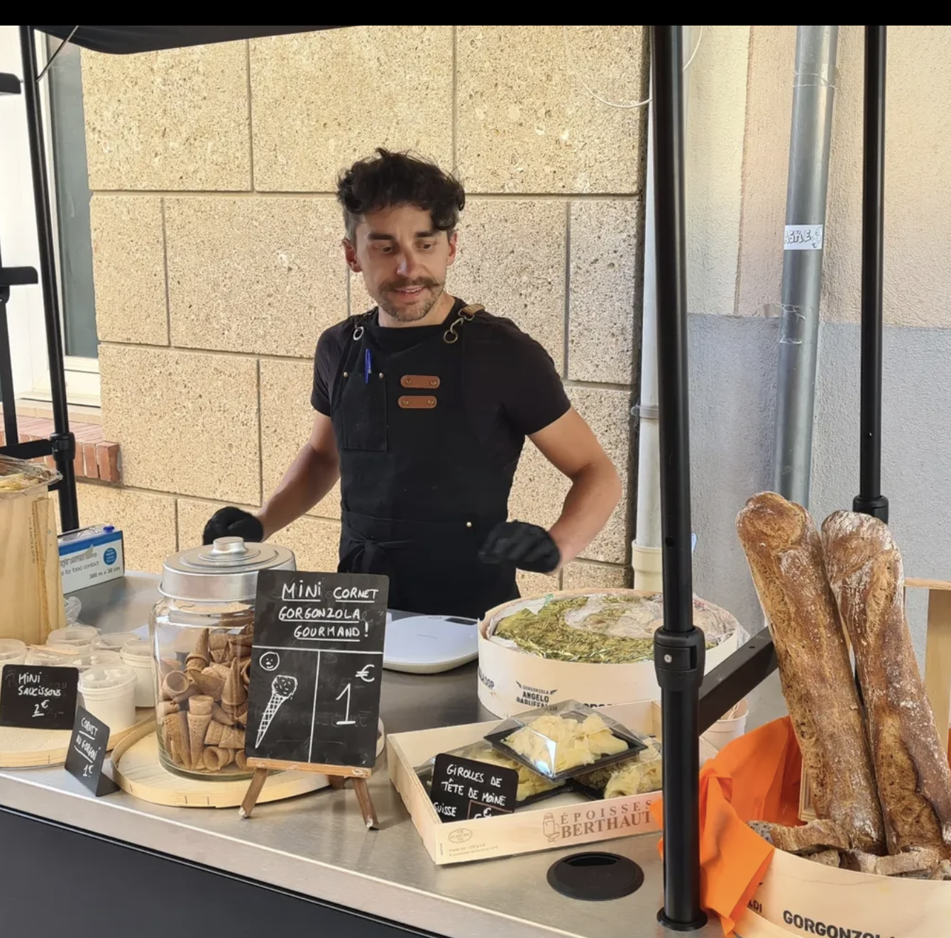 A man with curly dark hair and a beard, wearing a black apron and gloves, stands at a food stall selling baked goods. The stall has baguettes, ice cream cones, and various packaged treats, with signs in French indicating prices and items.