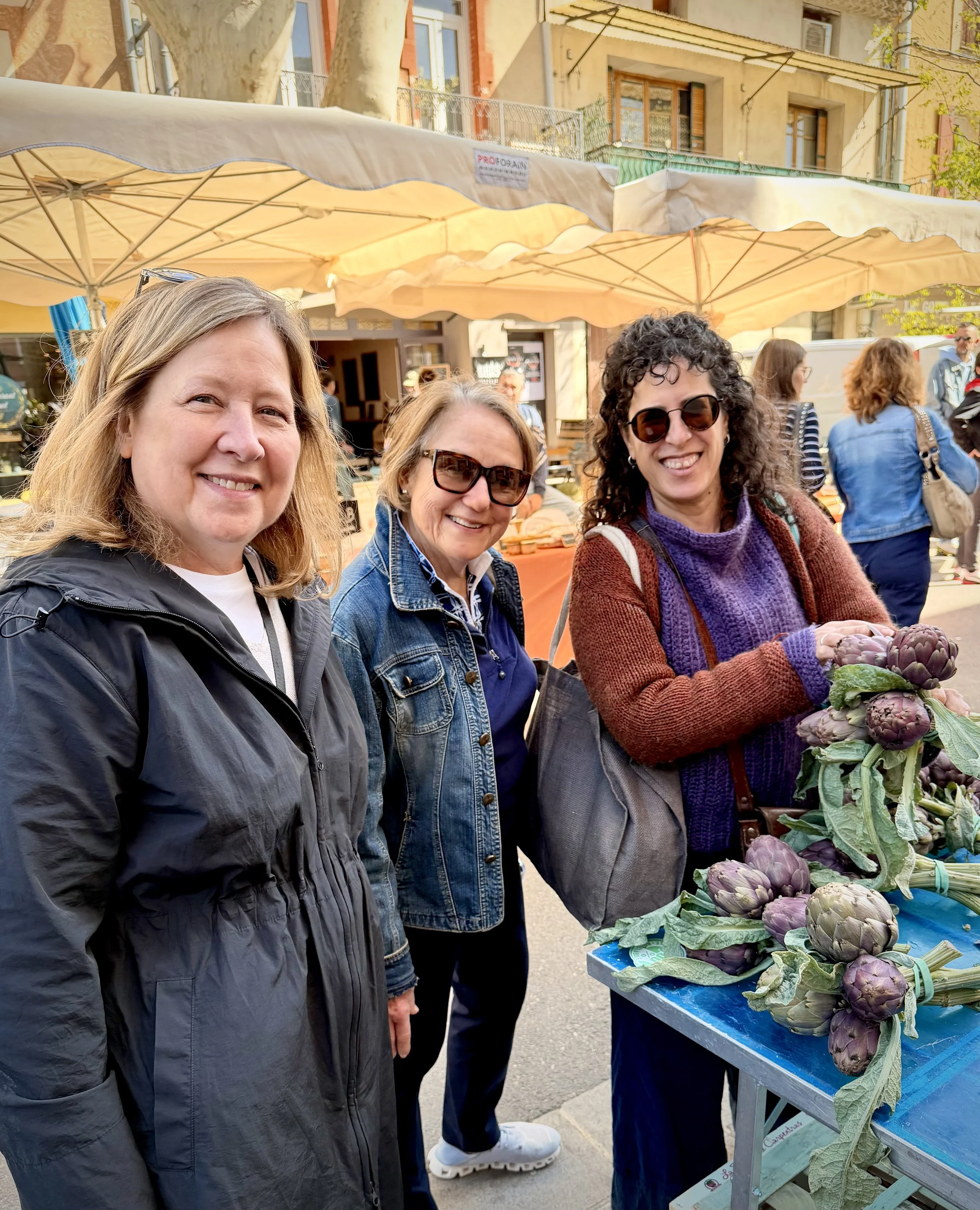 Three women standing at an outdoor market stall selling artichokes. They are smiling and wearing casual clothing, with sunglasses. The market is set up on a city street with buildings and trees in the background.