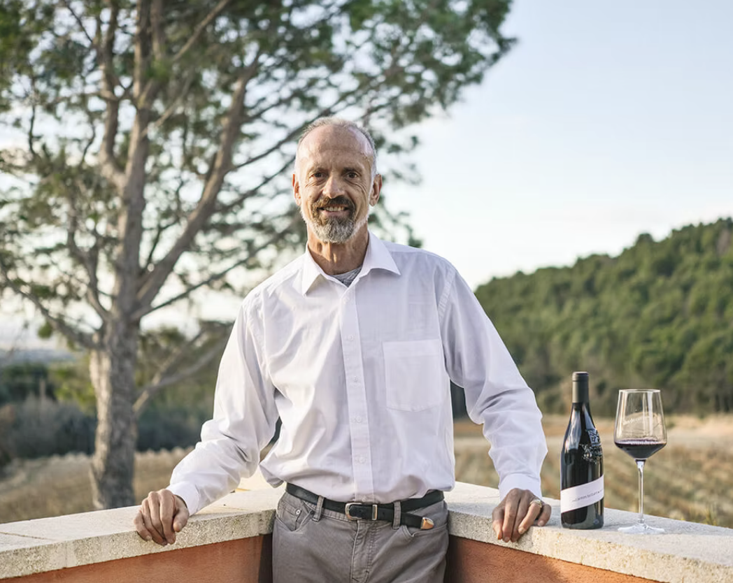 A middle-aged man in a white dress shirt standing on a balcony outdoors with a glass of red wine, a bottle of wine, trees, and a hillside in the background.
