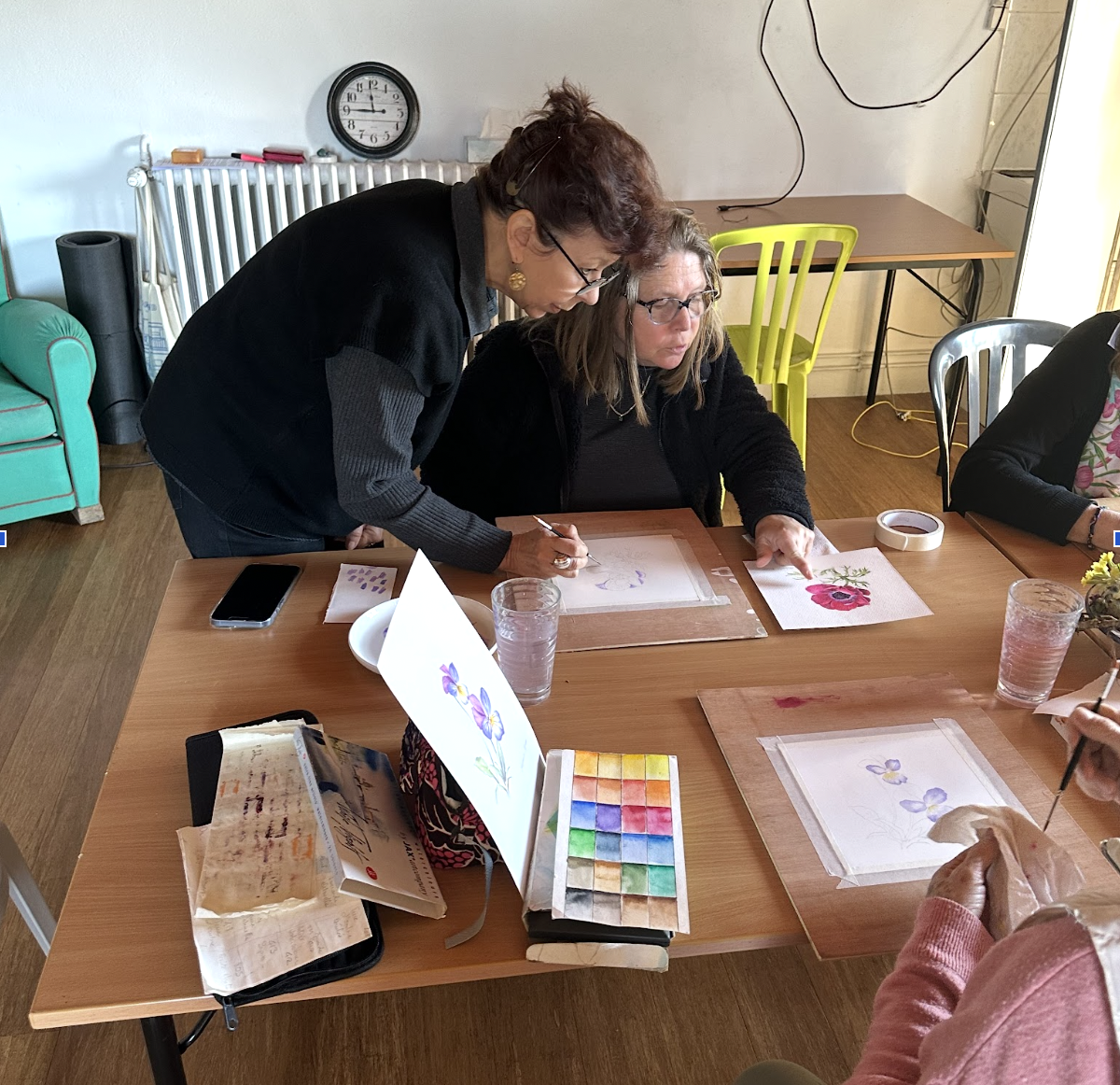 Two women engaged in watercolor painting at a wooden table, with art supplies such as watercolor paints, brushes, and paper with floral watercolors.