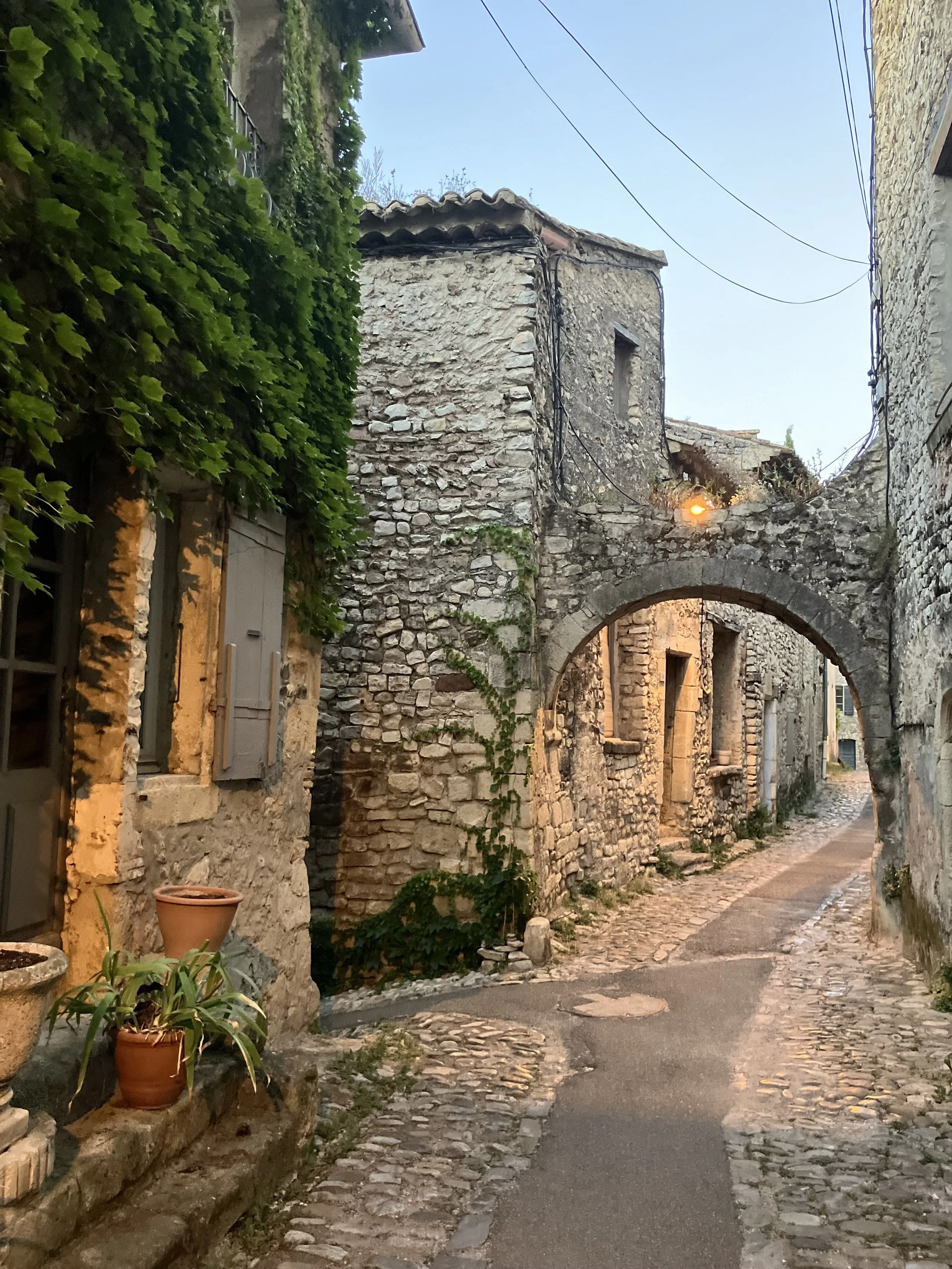 A narrow cobblestone street in an old European village with stone buildings, some with wooden shutters, and ivy climbing on the walls. The scene is lit by warm streetlights, with a clear evening sky above.