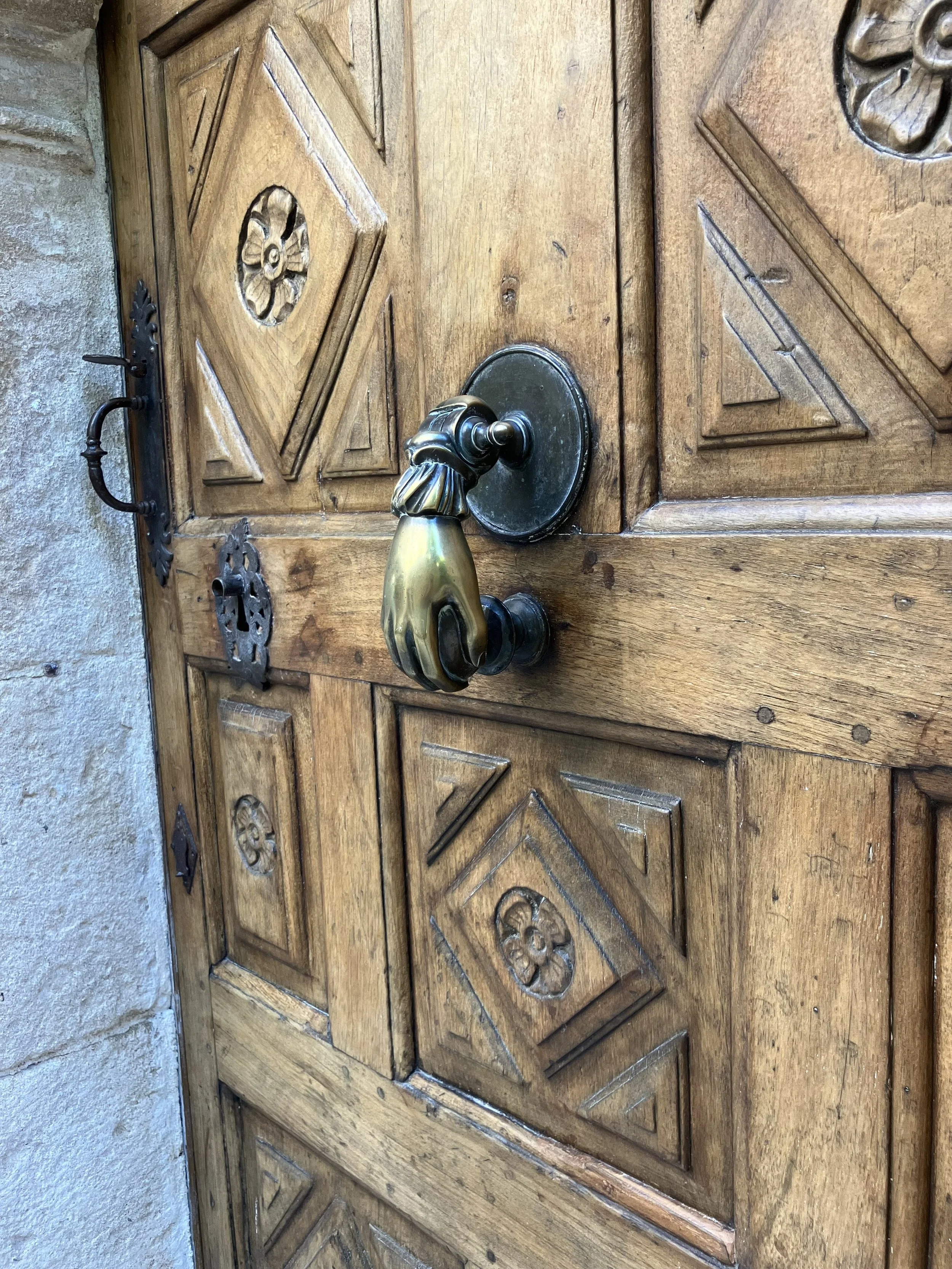 Close-up of a wooden door with decorative carved panels, black wrought iron hinges, and a brass door handle with a decorative backplate, set in a stone wall.