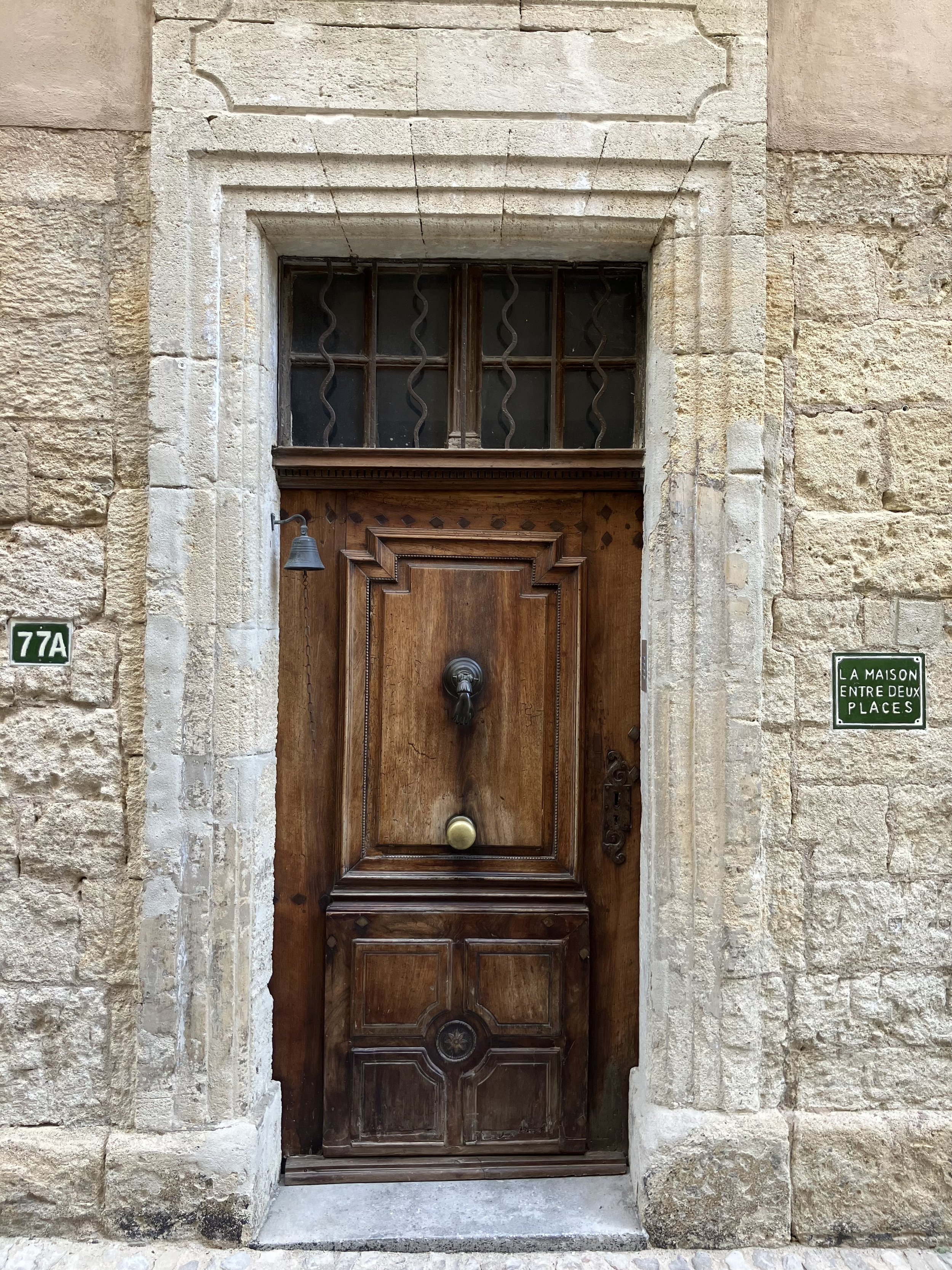 A wooden door with a decorative design in the center, set in a stone wall with a window above it featuring wrought iron bars. Two small green signs are on either side of the door, one reads '77A' and the other reads 'La maison entre deux places'.