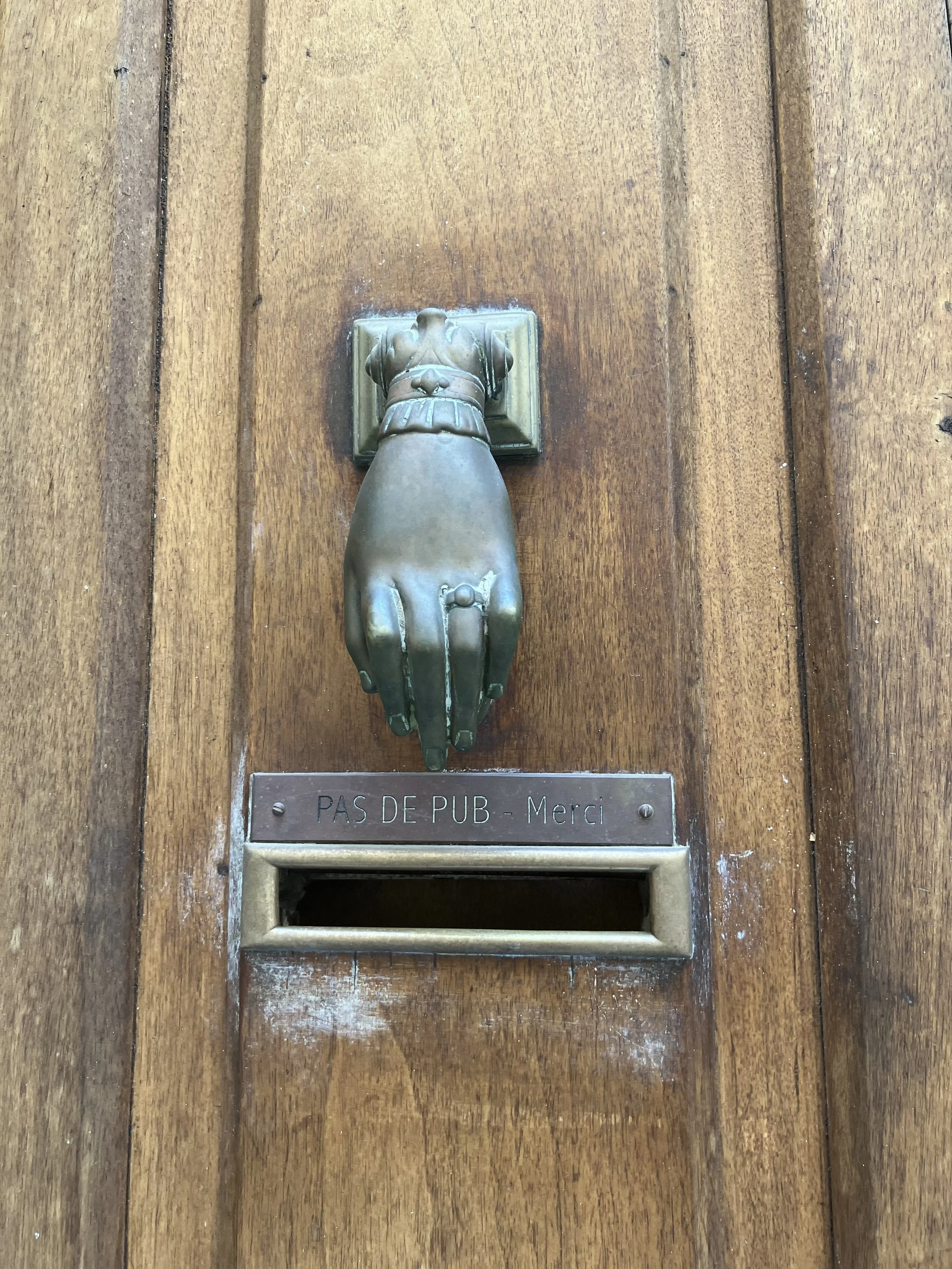 Wooden door with a metal door handle designed as a hand. There is a small rectangular slot below the handle. A sign below the slot reads "PAS DE PUB - Merci".