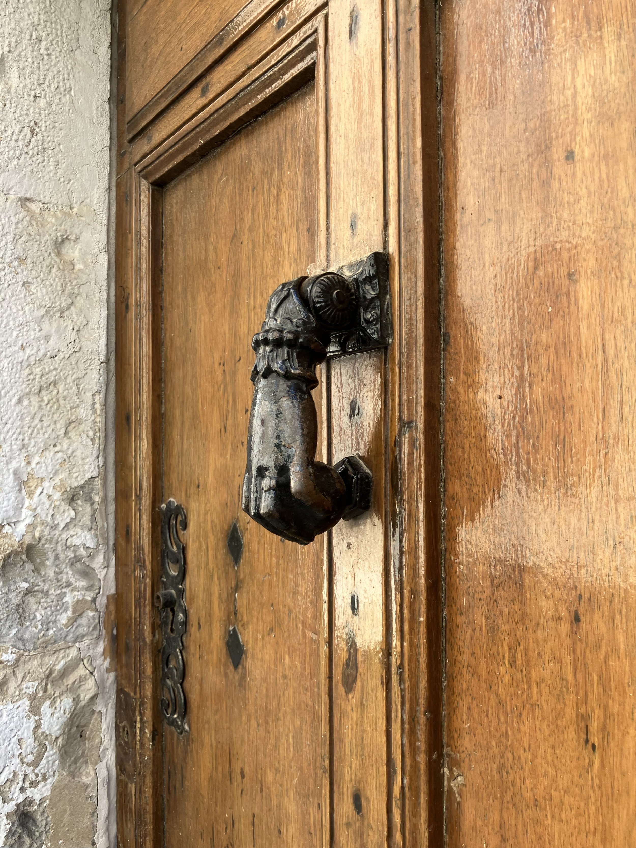 An ornately carved black door handle on a vintage wooden door, with a chain lock and metal hinges, against a stone wall.