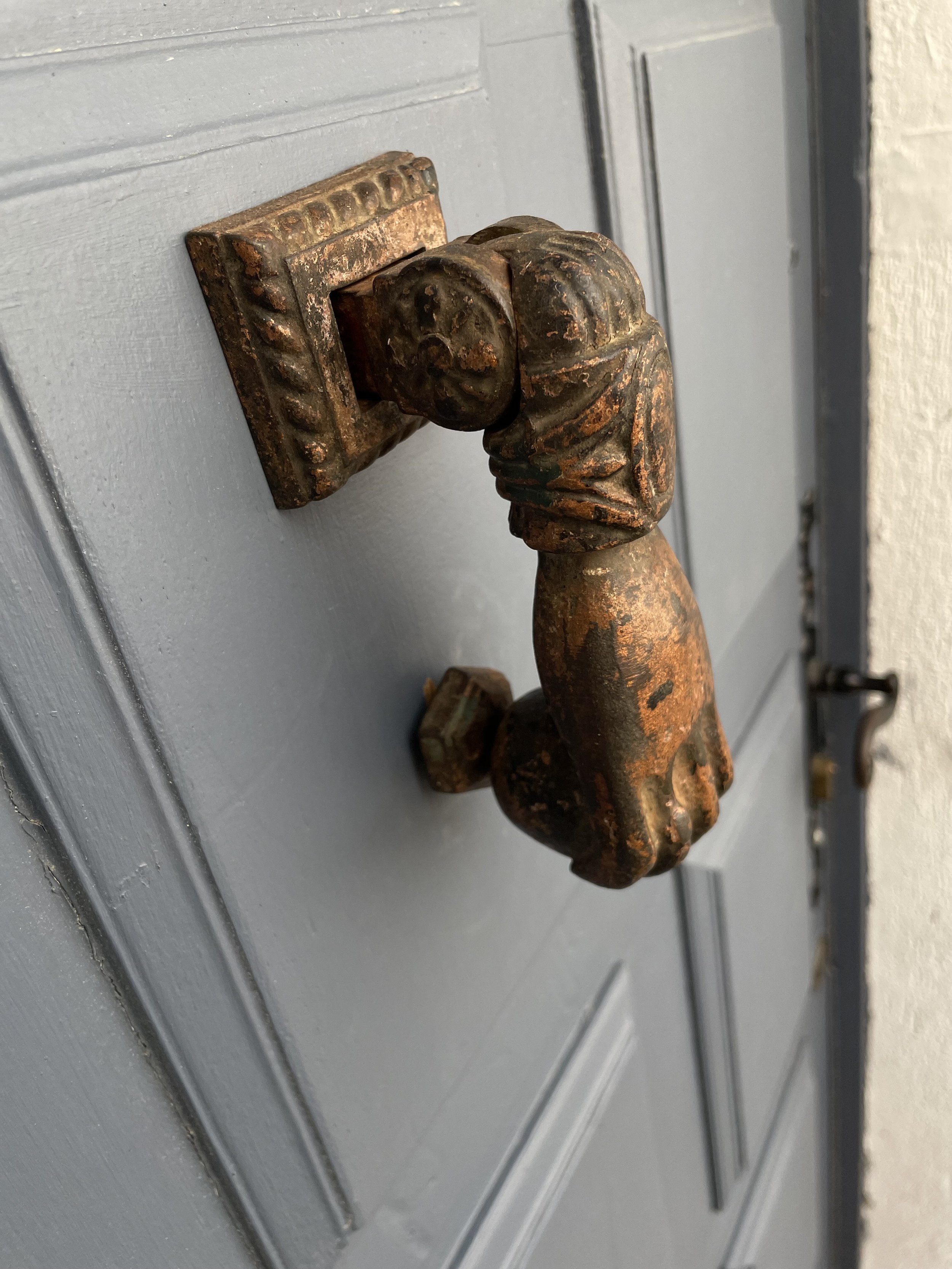 Close-up of a vintage, rusted door handle shaped like a clenched fist, mounted on a gray door.
