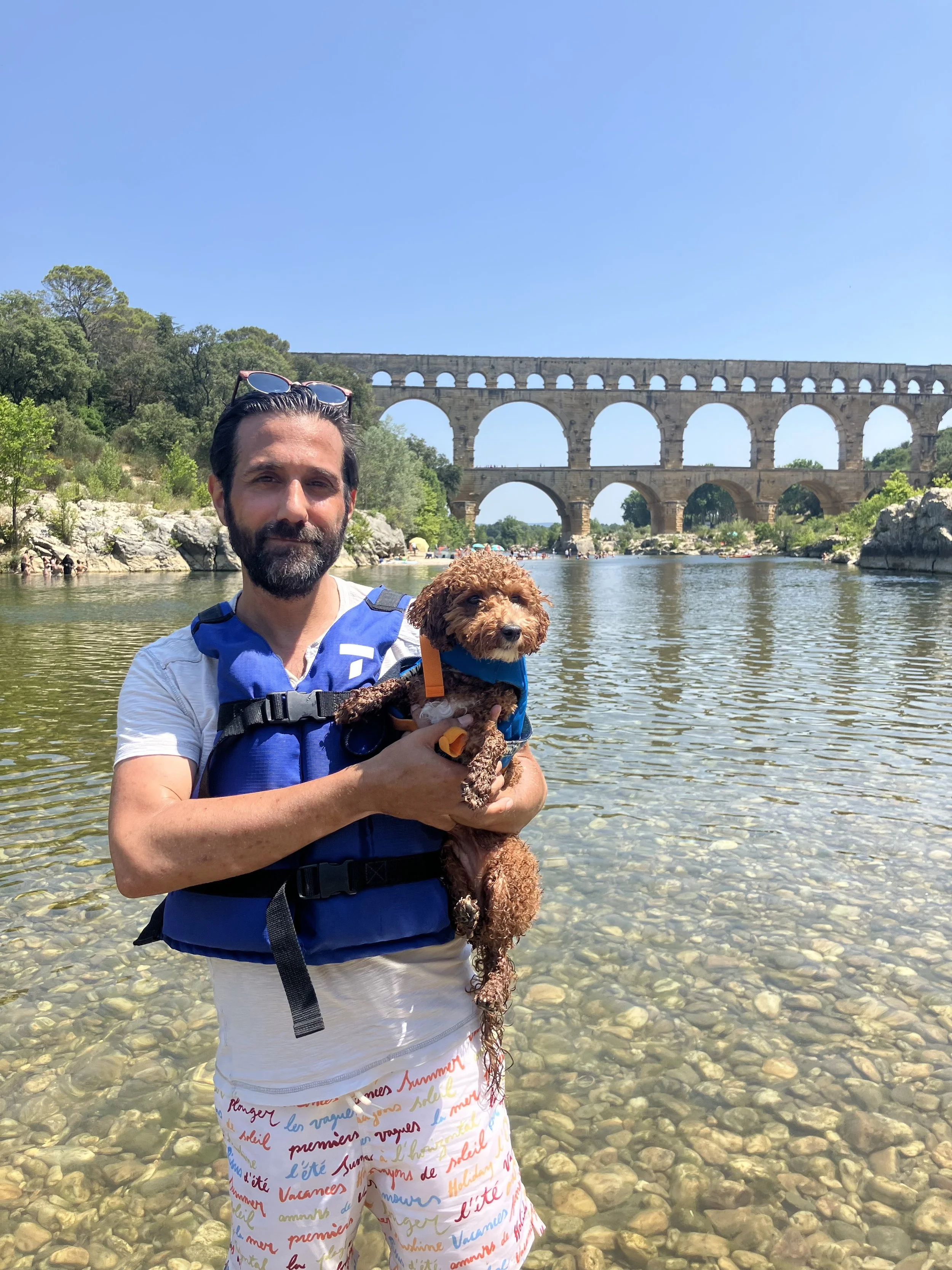 A man with a beard holding a small brown Poodle dog in both arms, standing in a river with a rocky bottom. The man is wearing a white shirt, colorful shorts, and a blue life jacket. In the background, there is the ancient Roman aqueduct Pont du Gard, and the sky is clear and blue.