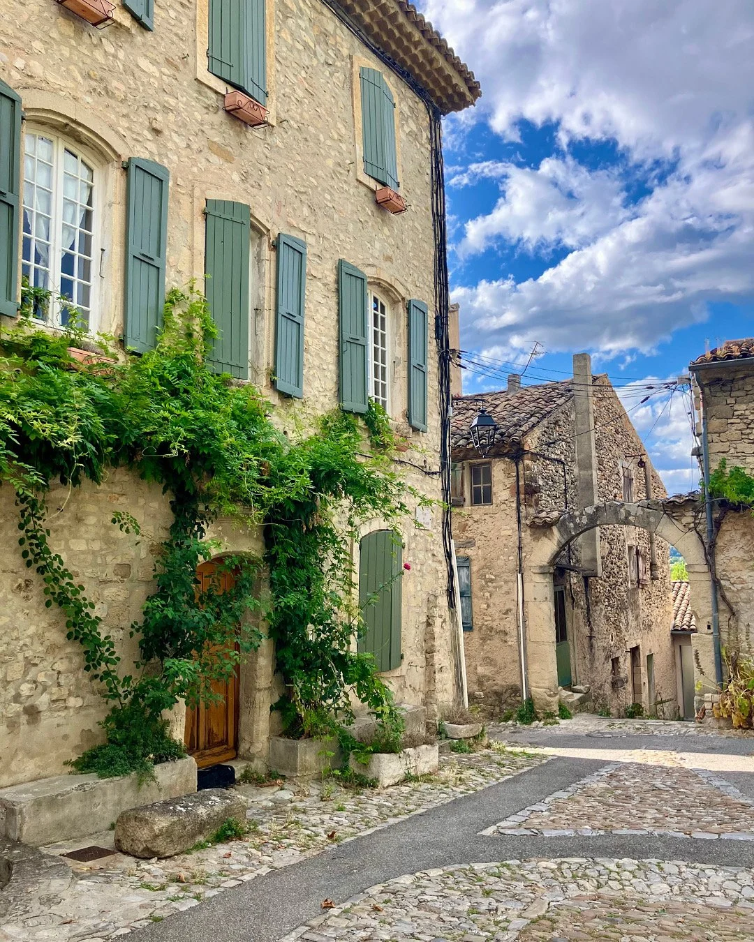 A cobblestone street in a historic European village with stone buildings, green shutters, and a stone archway against a partly cloudy sky.