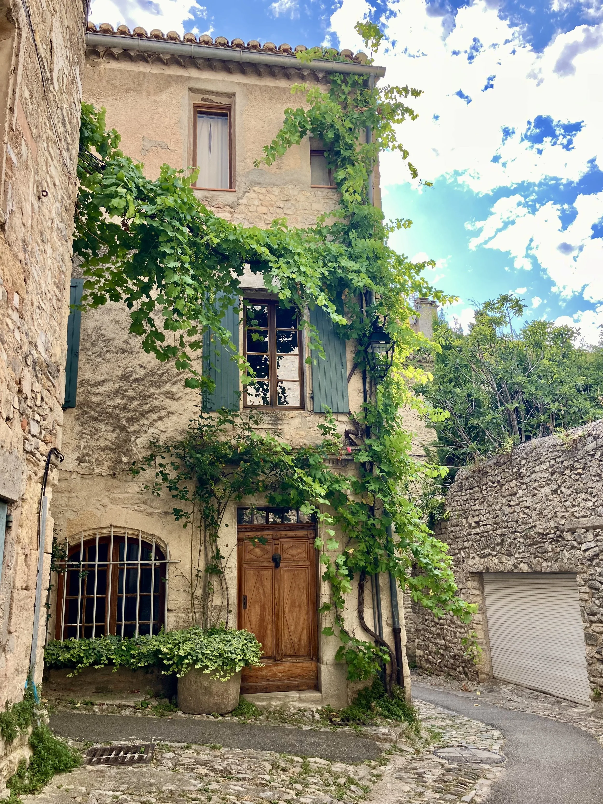 A rustic, multi-story stone house with wooden shutters and a vine growing over the windows and door, set in a narrow cobblestone street under a partly cloudy sky.
