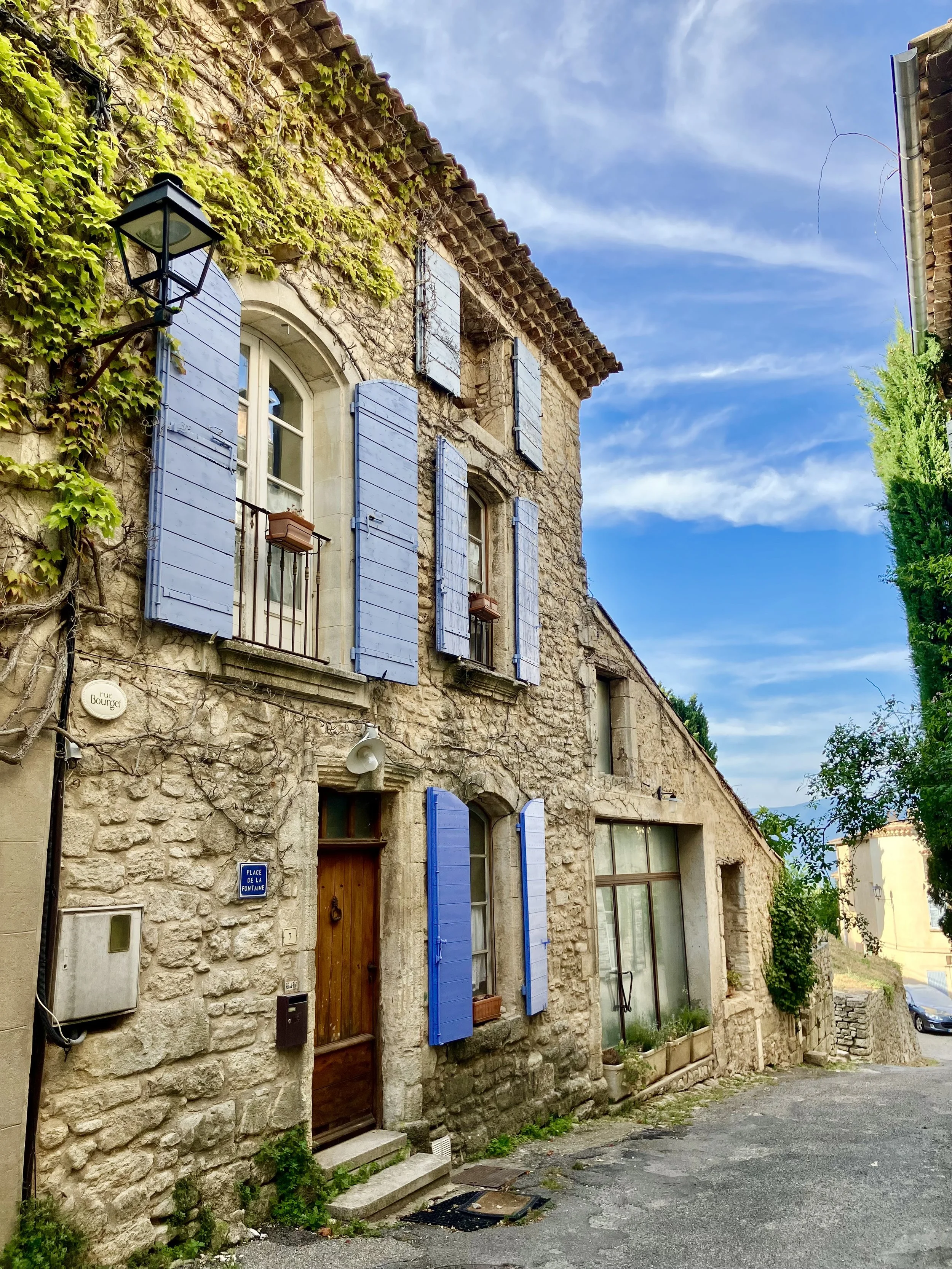 A stone building with blue shutters and a wooden door on a narrow street, ivy growing on the wall, and a street lamp, under a partly cloudy blue sky.