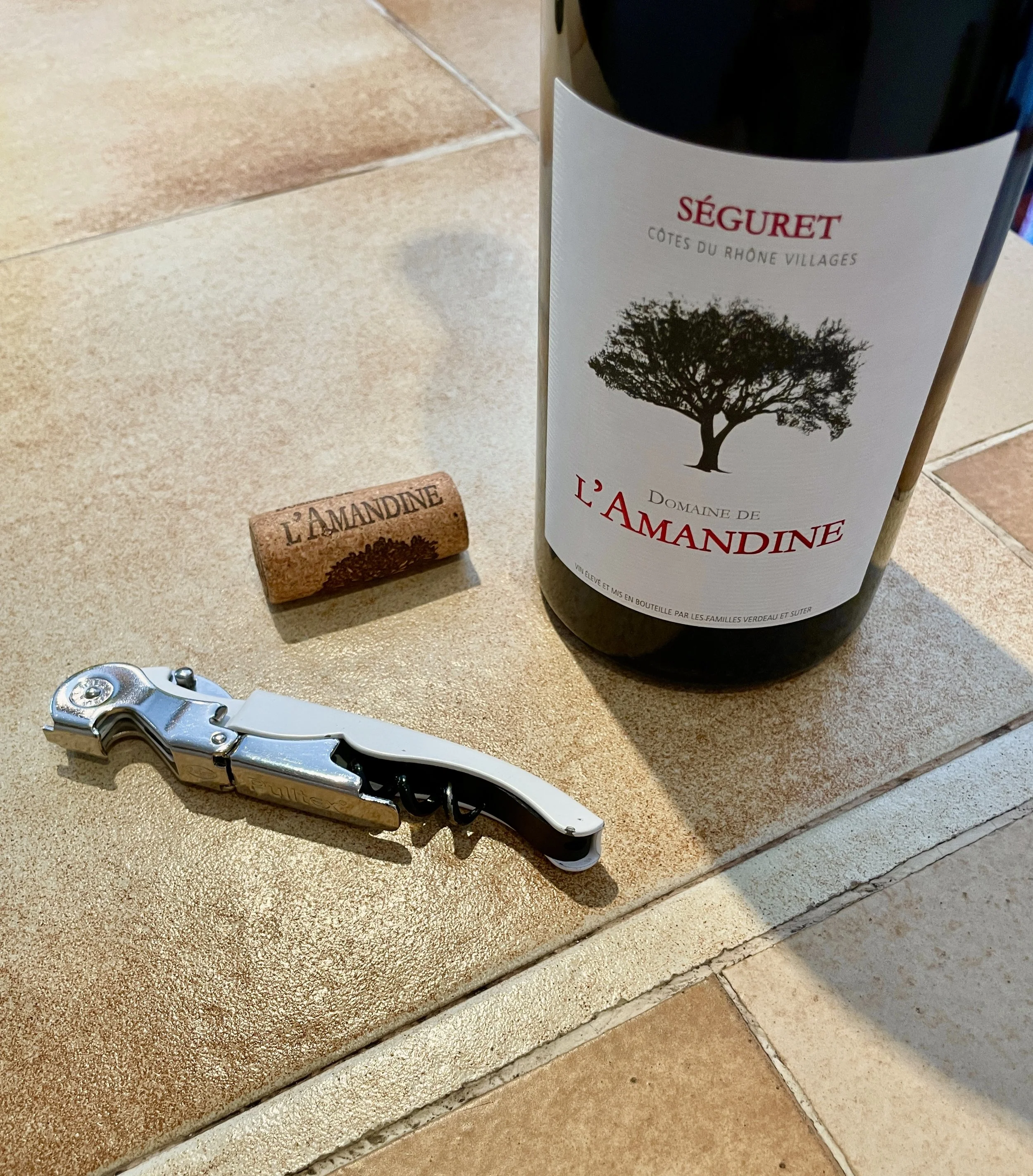 A wine bottle with a label indicating Domaine de L'Amandine, a cork with the same branding, and a corkscrew on a beige tiled surface.