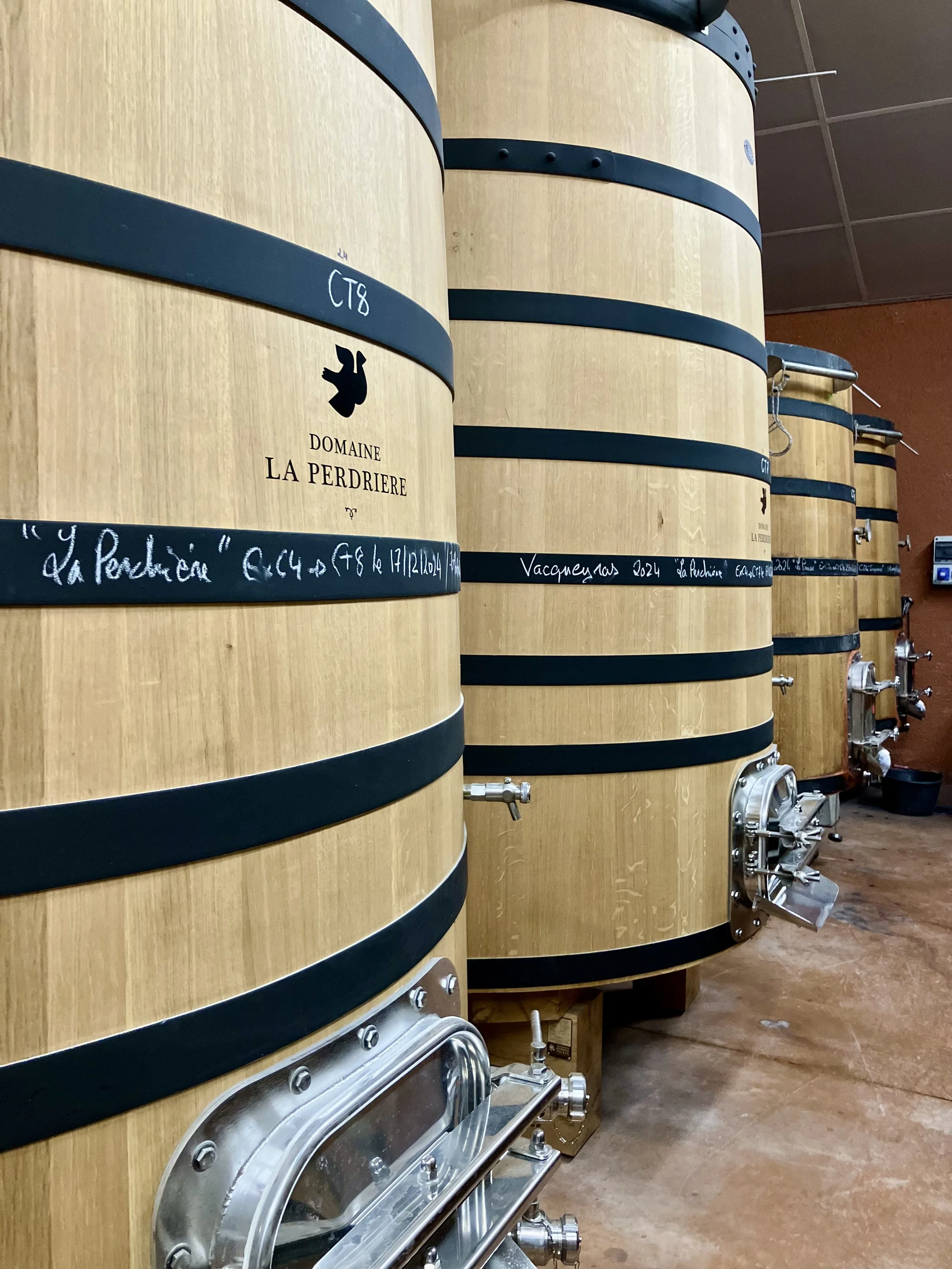 Several large wooden barrels with black bands, metal spigots, and handwritten labels, positioned on a wooden floor in a wine cellar or storage room.