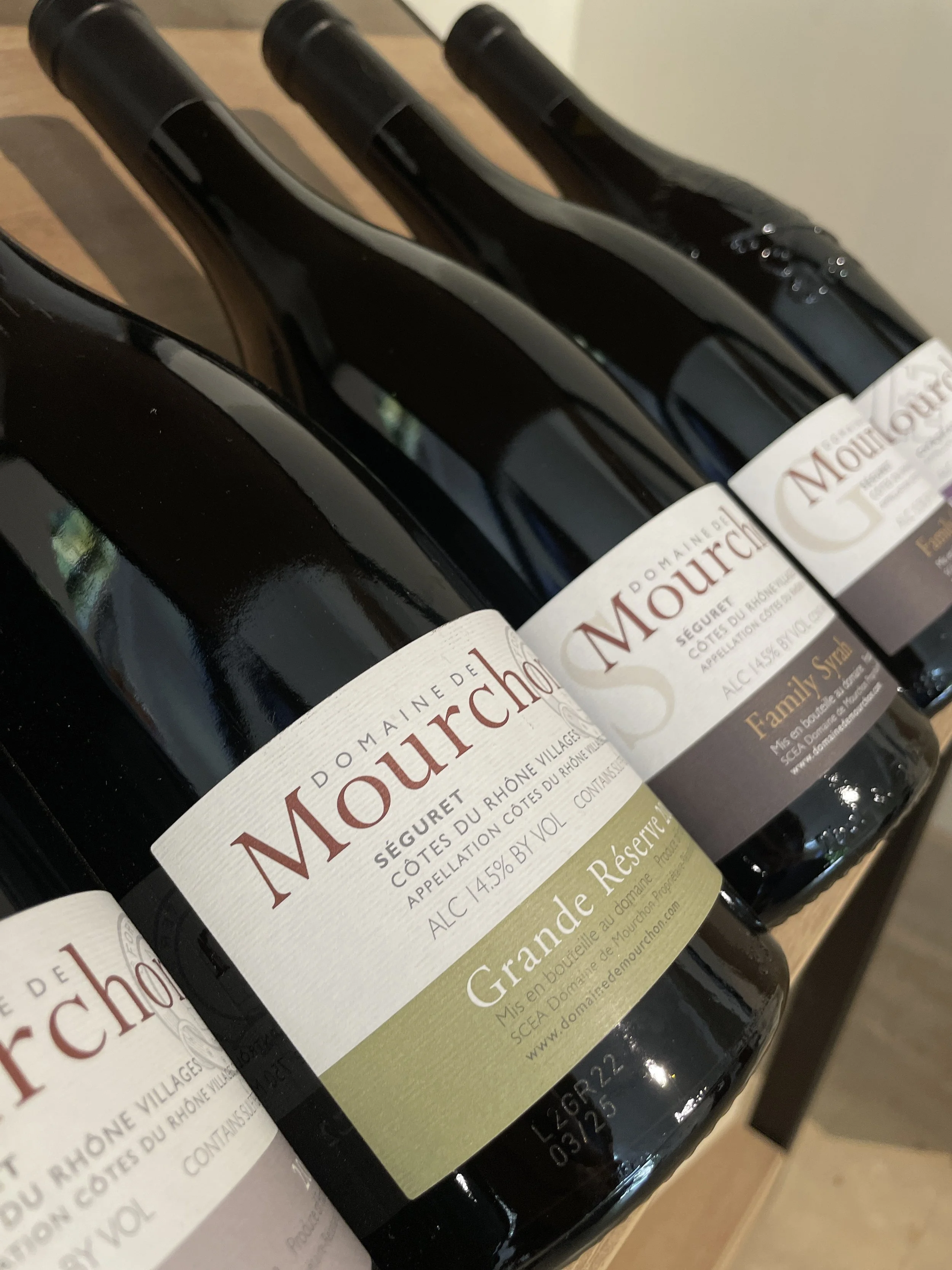 Close-up of three bottles of Domaine de Mourchon red wine, laying horizontally on a wooden surface with labels visible, showing the wine's name, vintage, and alcohol content.