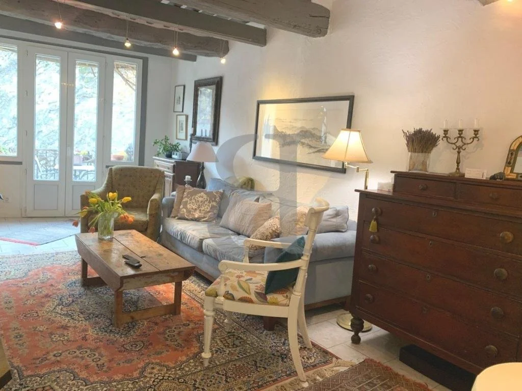 Living room with large glass doors, a wooden coffee table with a flower vase, a beige armchair, a blue sofa with patterned cushions, a white armchair, a wooden dresser with a candle holder and dried flowers, side lamps, framed artwork on the wall, and decorative objects.