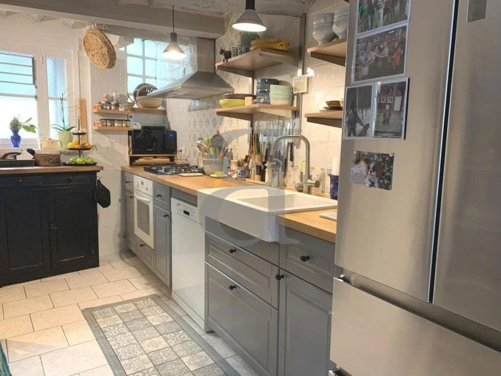 Kitchen with light gray lower cabinets, black upper cabinet, open wooden shelves, stainless steel refrigerator, stove, cutting board, various bowls, utensils, and decorative items, two windows with plants, and hanging pendant lights.