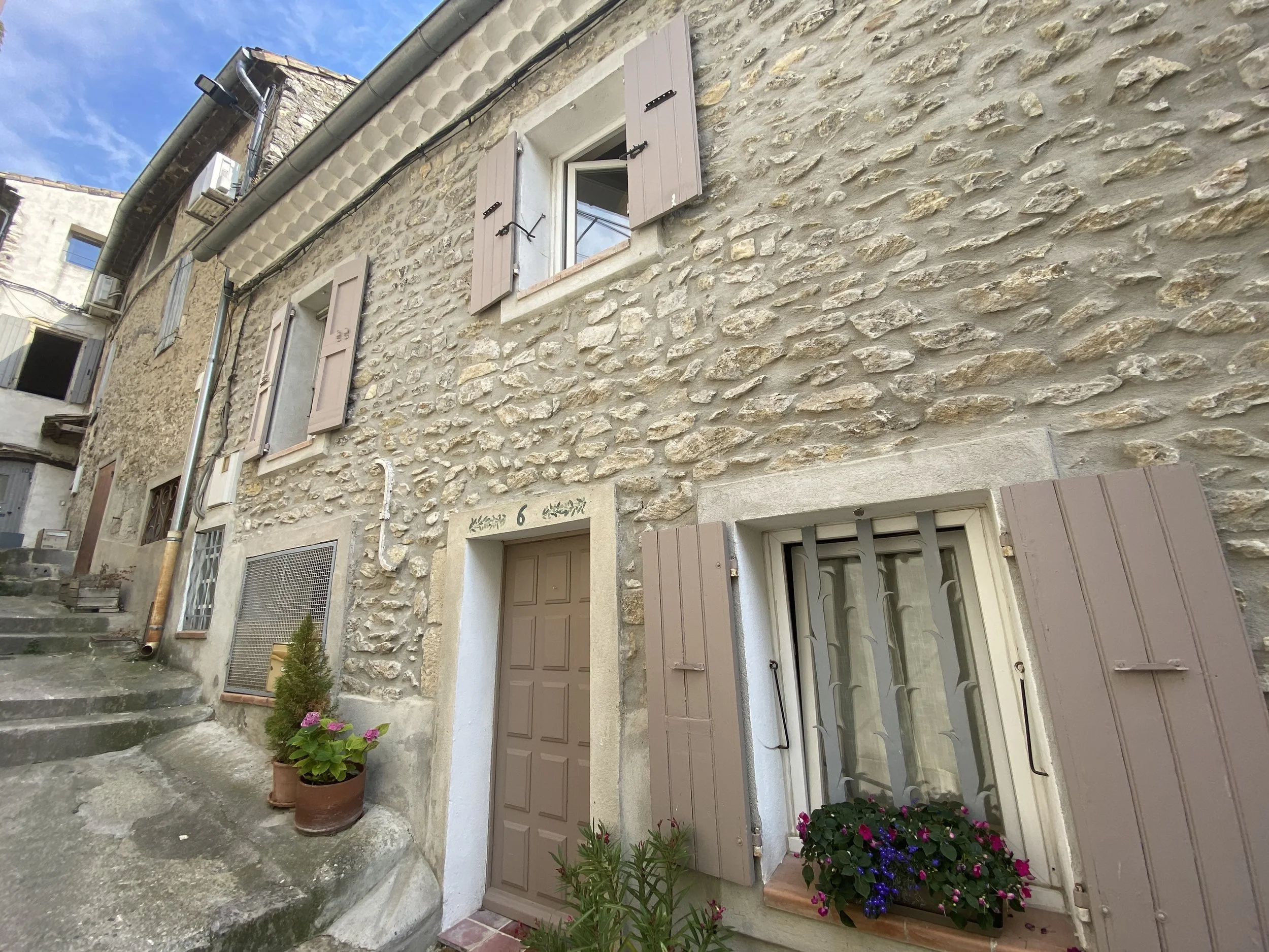 A stone house with closed beige window shutters and a beige door, small flower pots with colorful flowers, a small green shrub, stone steps, and a chimney against a partly cloudy sky.