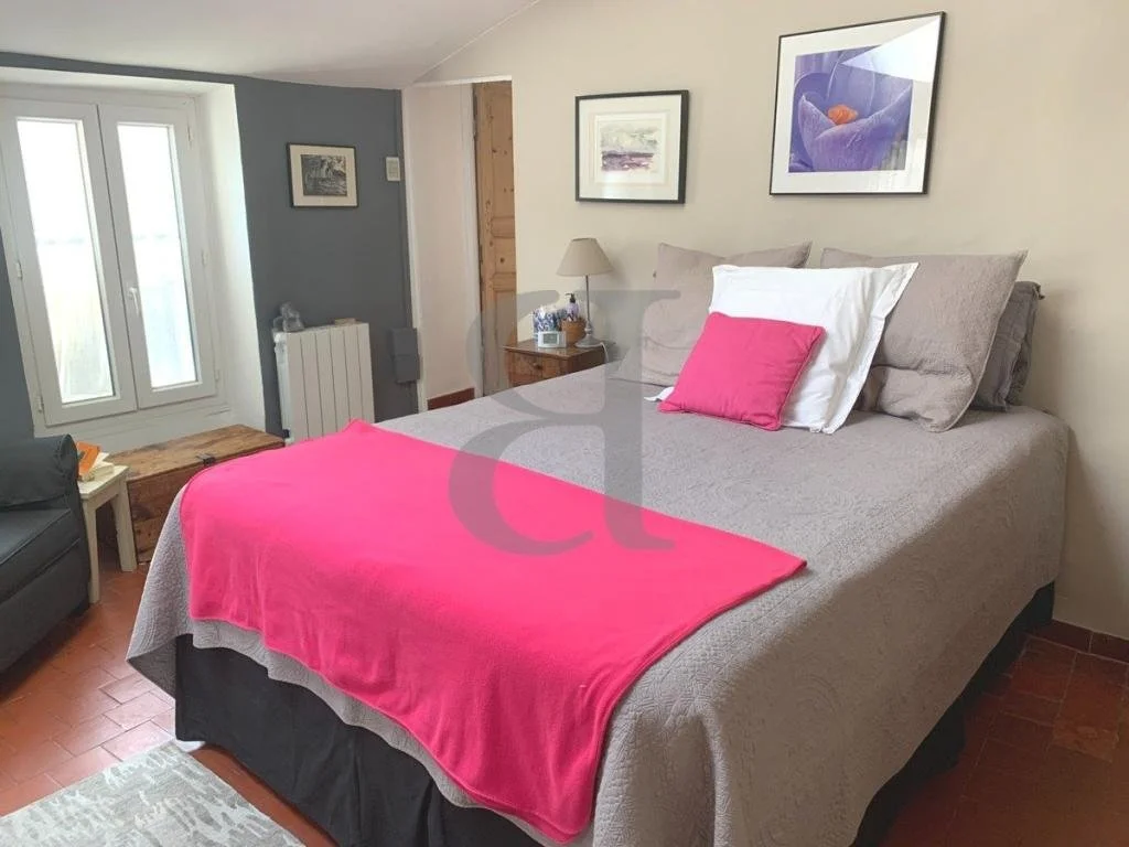 A cozy bedroom with a bed topped with gray and white pillows and a bright pink blanket, small wooden nightstand with a lamp, framed artwork on the cream walls, and French doors letting in natural light.