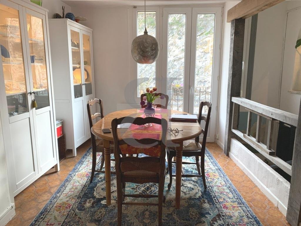 A cozy dining room with a wooden table, four chairs, a hanging pendant light, and a vase with tulips on the table. There are white cabinets to the left, large windows and a glass door letting in natural light, and a fireplace on the right with a wooden mantel. An ornate rug covers the floor.