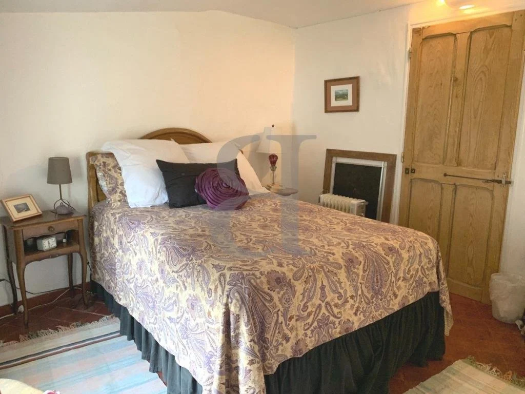 A bedroom with a bed covered in a paisley-patterned bedspread, several pillows, and a wooden headboard. To the side, a nightstand with a lamp, picture frame, and alarm clock. On the wall, there's a framed picture. Next to the window, a small mirror or picture frame leans against the wall, and below it is a radiator. Wooden double doors are closed, and a small wastebasket is on the floor.