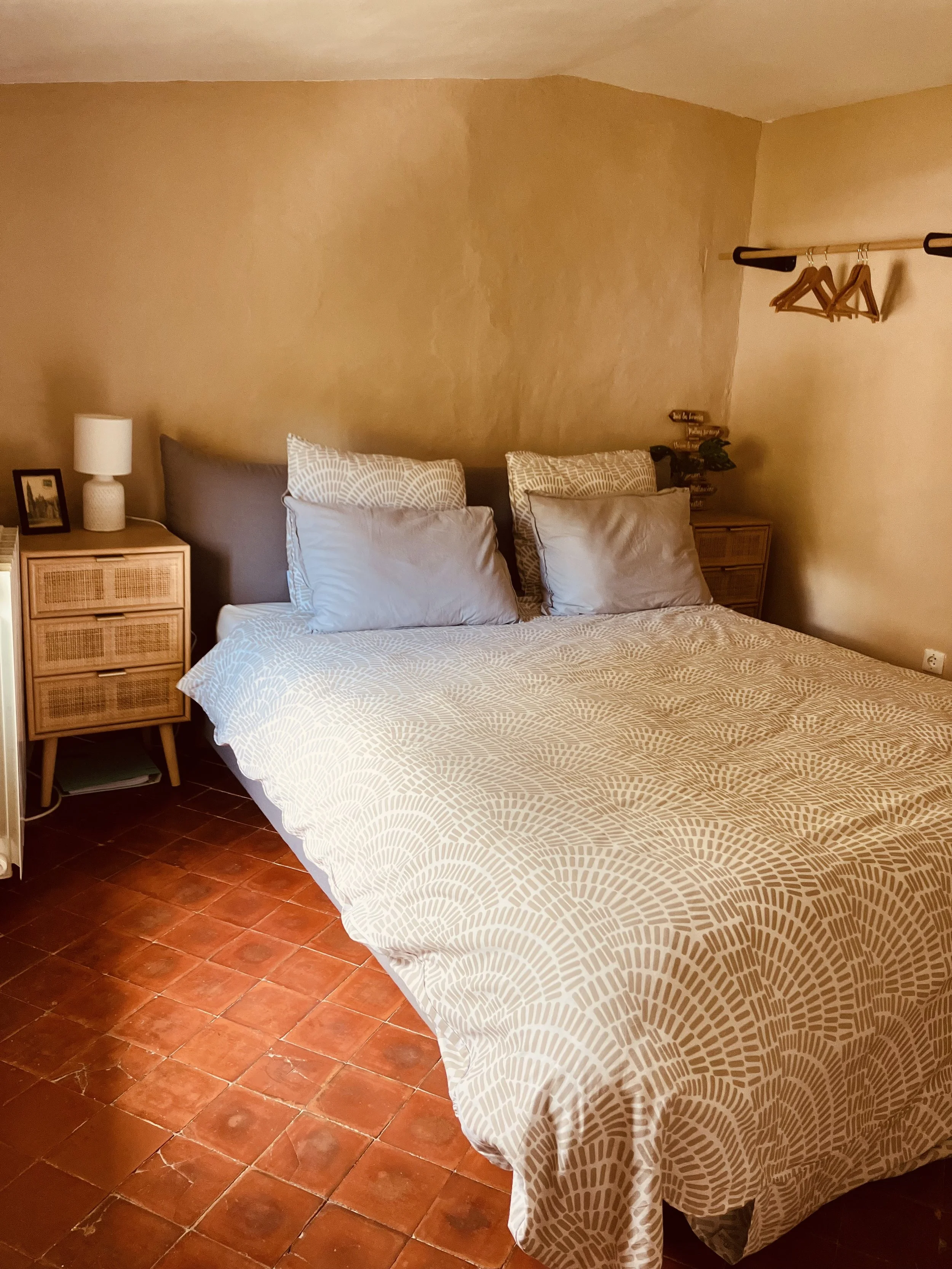 A bedroom with a bed, pillows, nightstand, lamp, and decorative items on the wall. The room has terracotta tile flooring and beige walls.