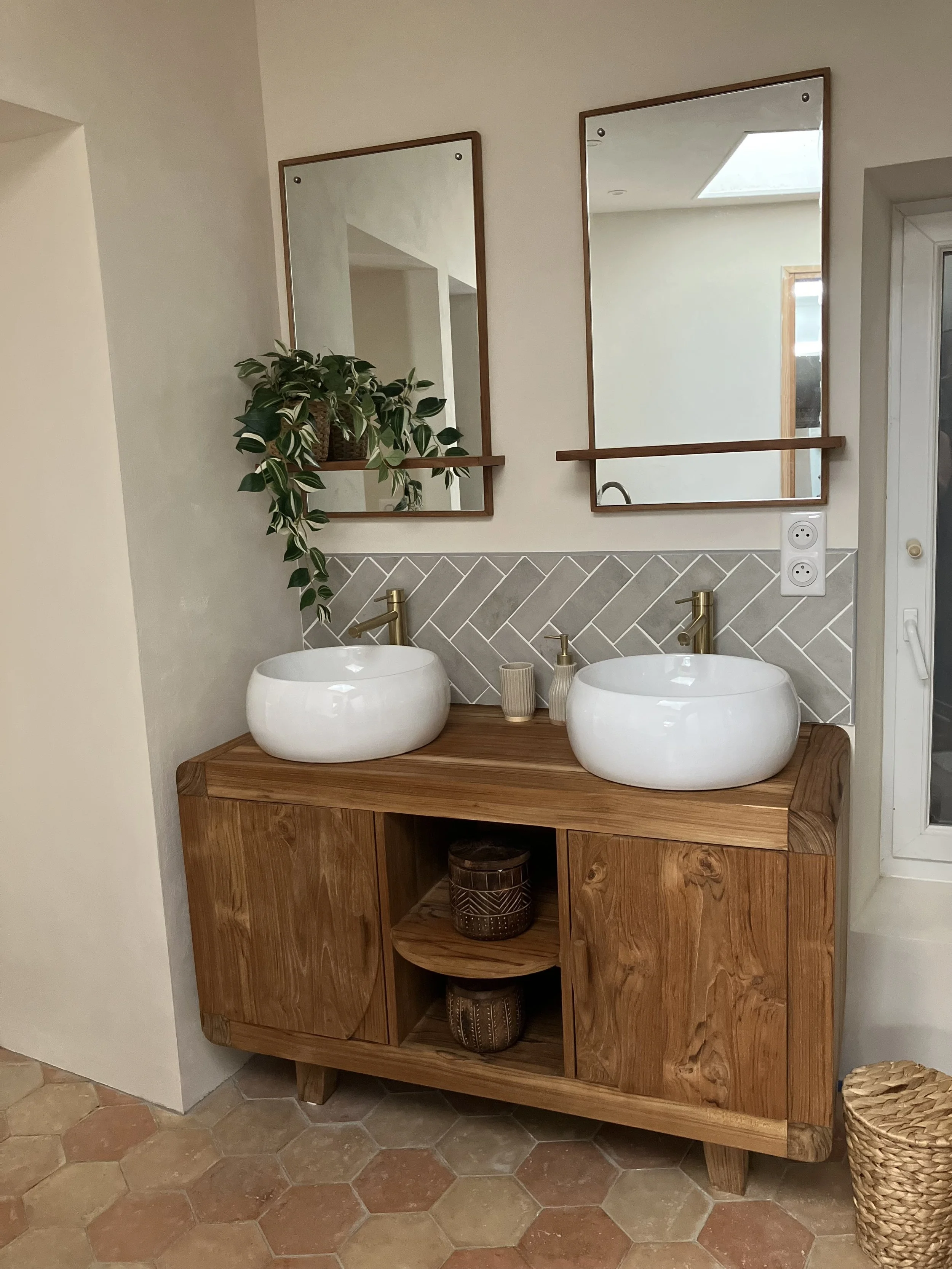 A bathroom vanity with two round vessel sinks, gold faucets, and two large rectangular mirrors. There is a potted plant and decorative items on the vanity, with hexagonal tile flooring.