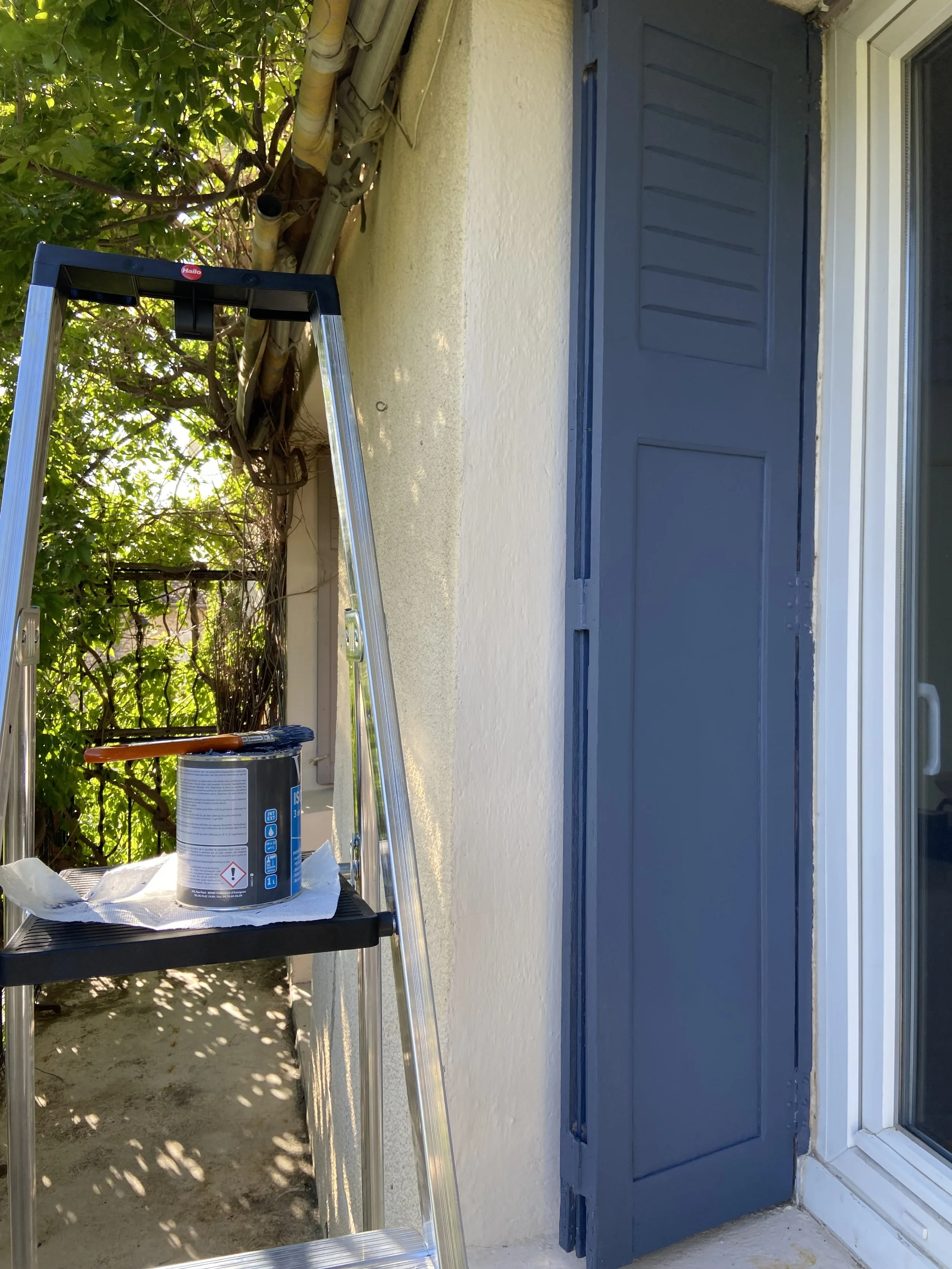 Photo of a small outdoor balcony or patio area with a silver step ladder, a can of paint, and some brushes placed on a black shelf. The background features a white wall, a blue shuttered window, and a tree with green leaves casting shadow.