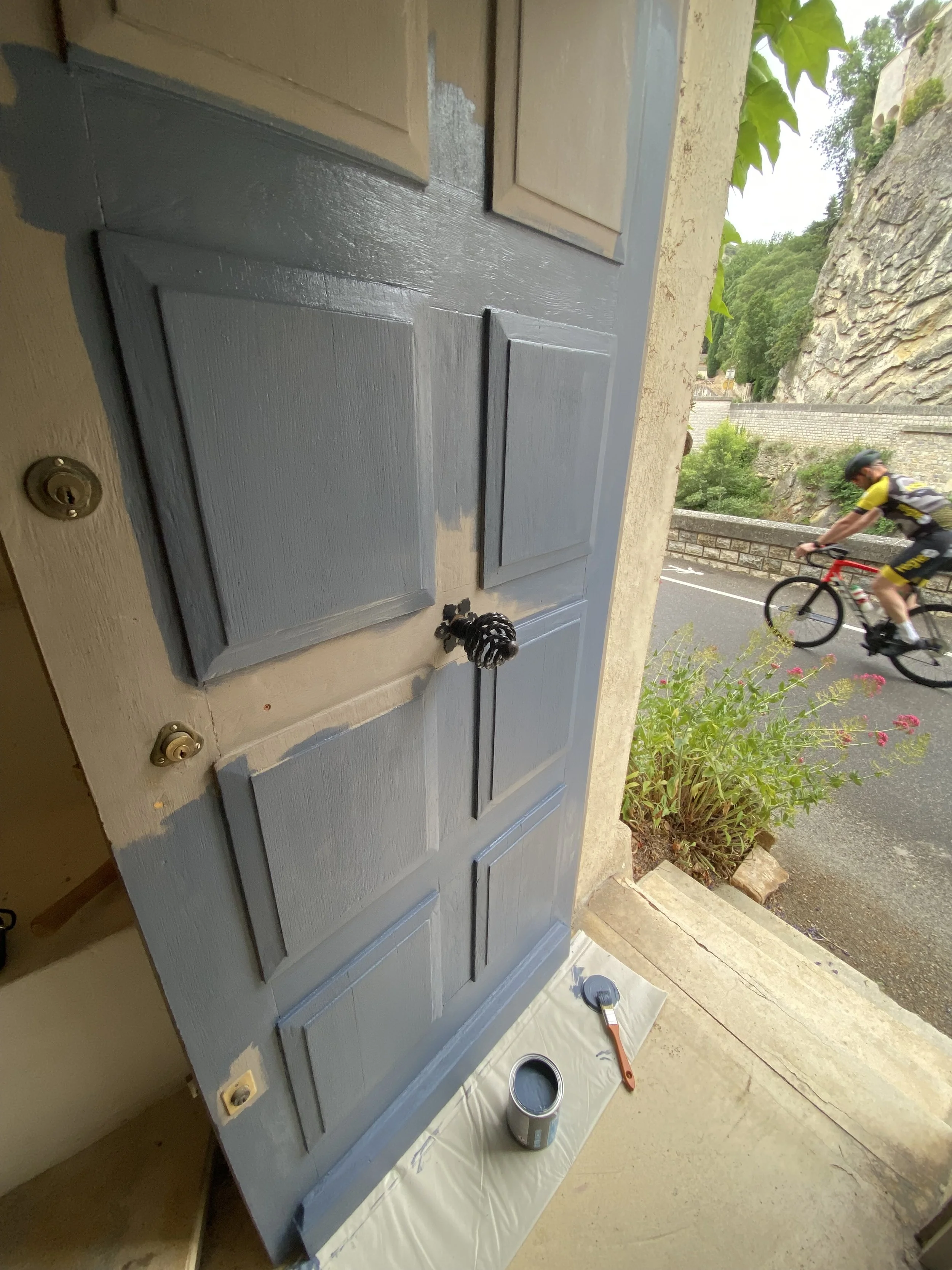 A house door is being painted with light blue paint. There are painting supplies on the ground including an open can of blue paint, a paintbrush, and painter's paper. Outside the door, a man wearing a black helmet and cycling gear is riding a bicycle