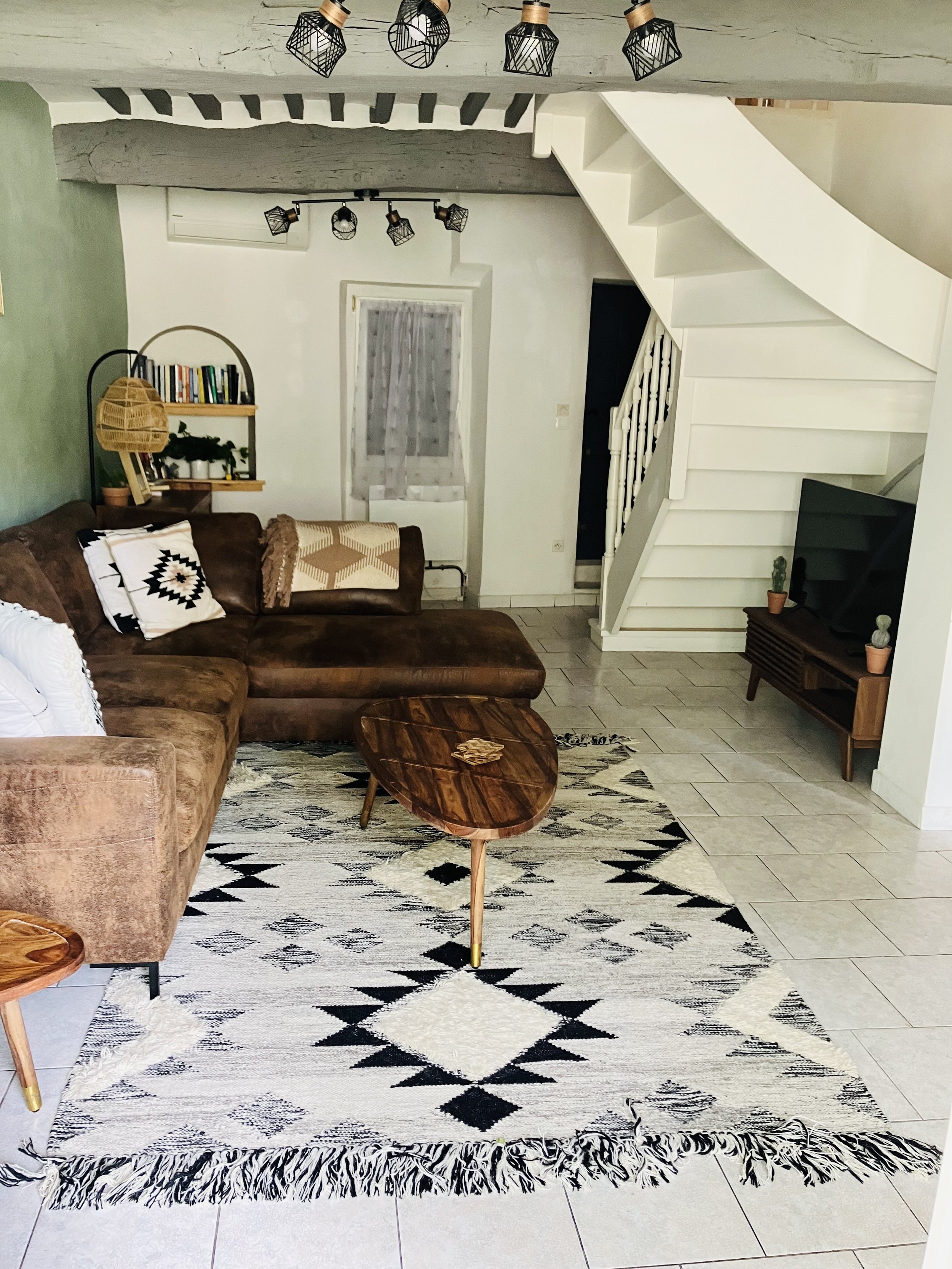 Living room with a patterned black and white rug, wooden furniture, a brown sectional sofa, a side table with small potted plants, wall-mounted bookshelf, white staircase, and ceiling light fixtures with open black wireframe shades.