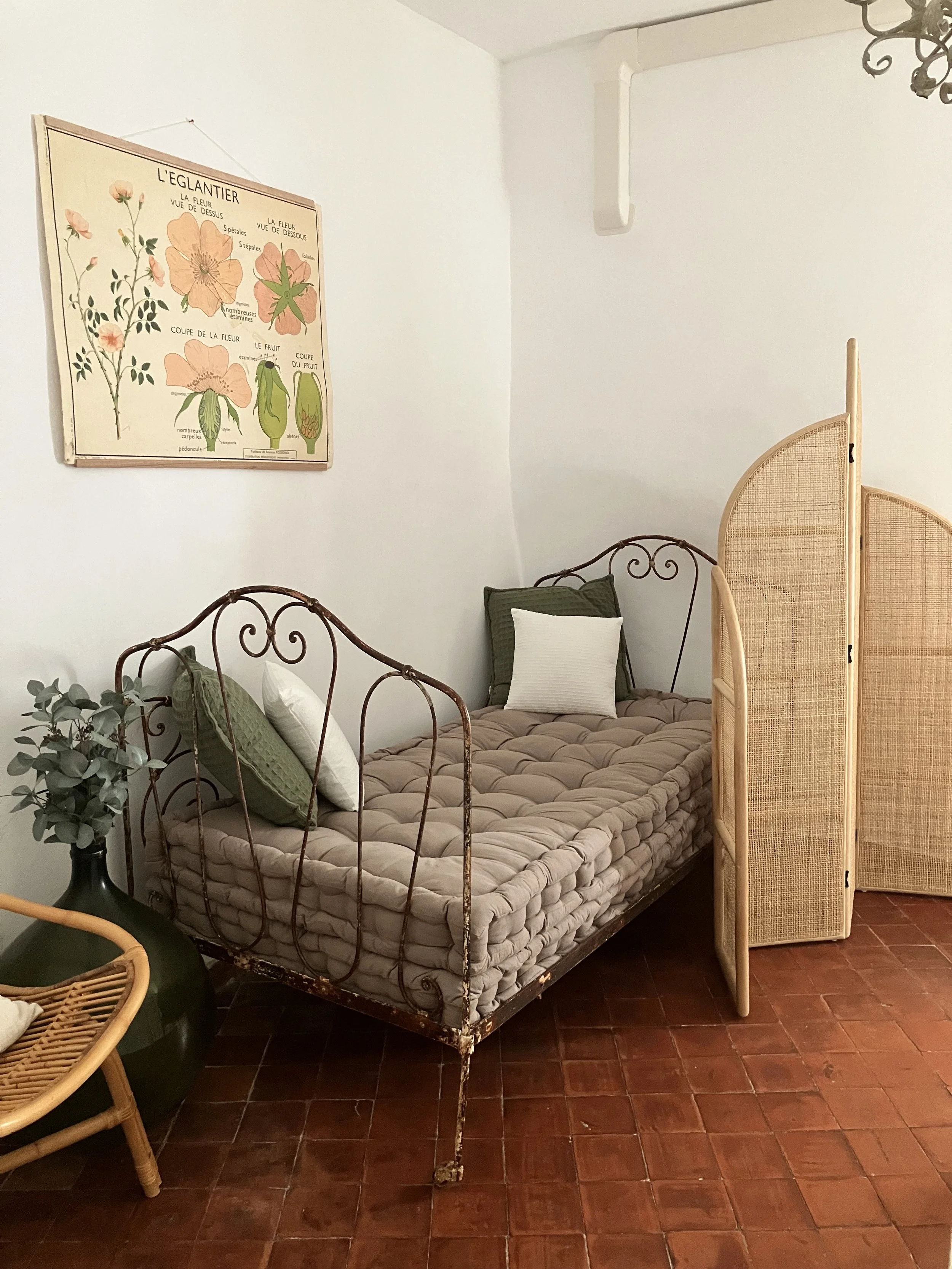 A room with a vintage iron bedframe with a tufted mattress, decorated with green and white pillows. Next to the bed is a large dark green vase with eucalyptus branches. Several wooden room dividers with woven panels partially block the view of the be