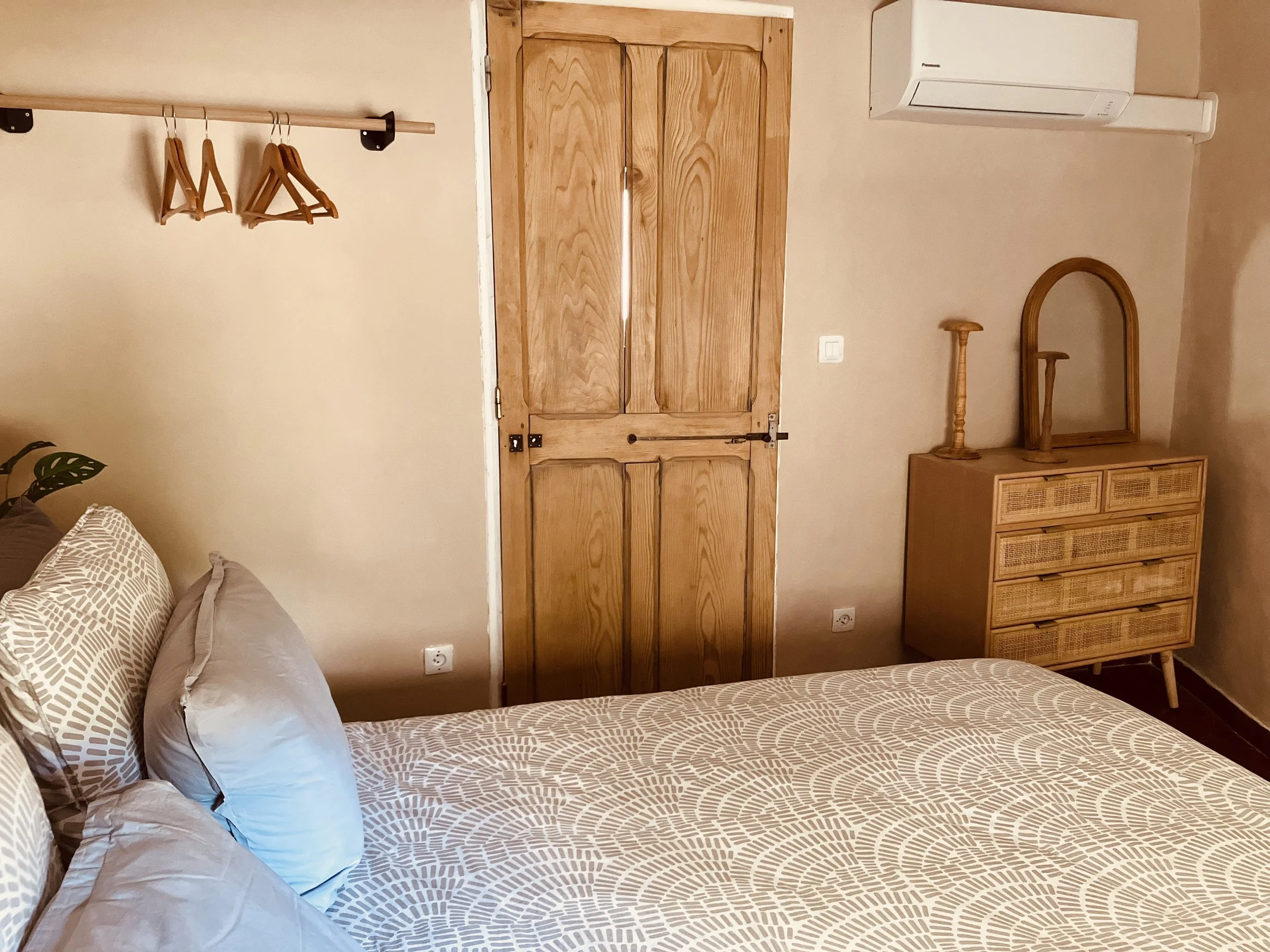 A bedroom with a bed in the lower left corner covered with a beige patterned bedspread, two pillows on the bed, a wooden wardrobe with double doors in the center, a rack with wooden hangers on the left, a wooden dresser with a mirror and two wooden d