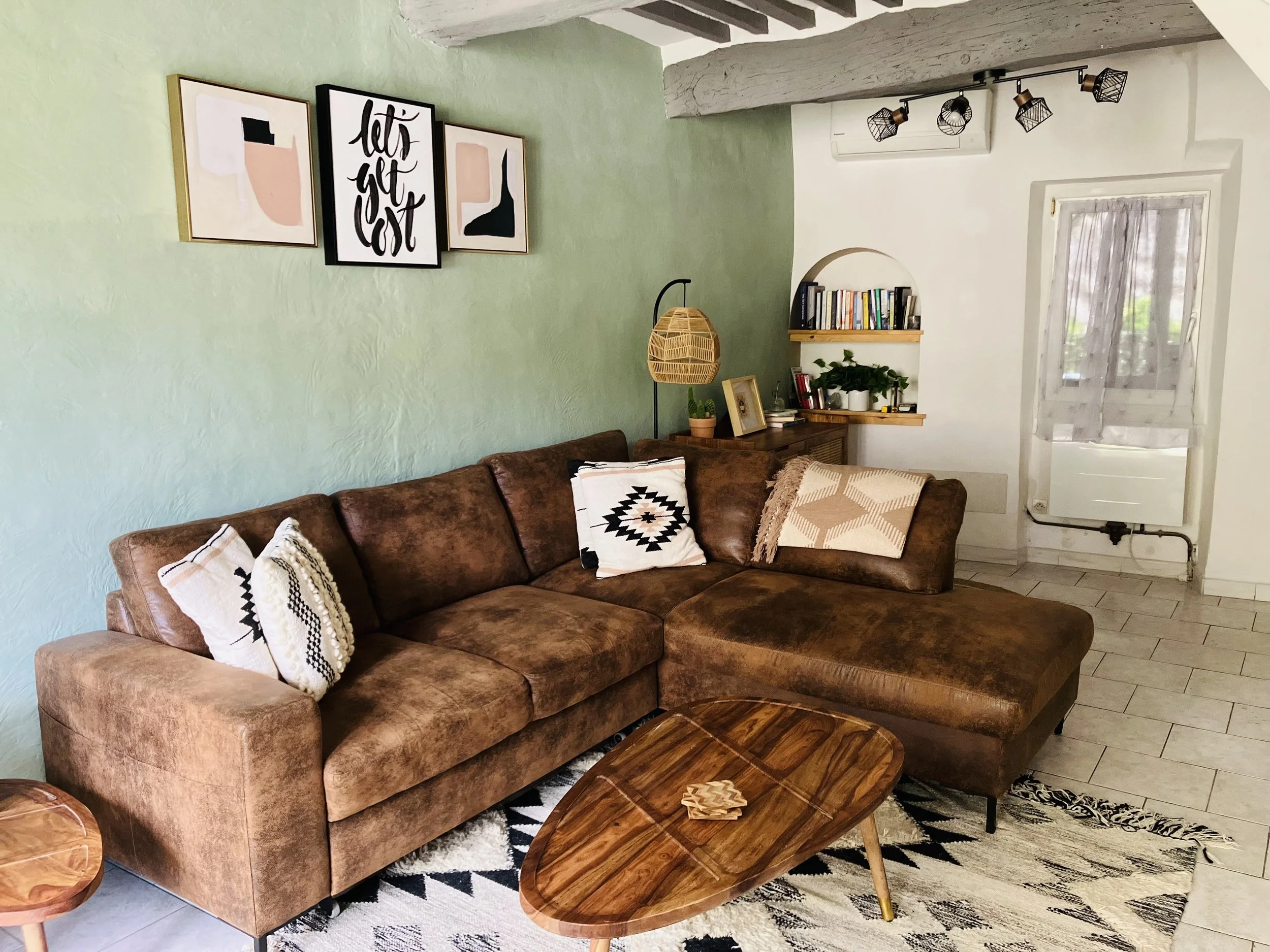 Living room with green textured wall, art prints, a brown leather sofa with patterned pillows, wooden coffee table, a bookshelf with books and plants, a window with curtains, and a track light fixture.