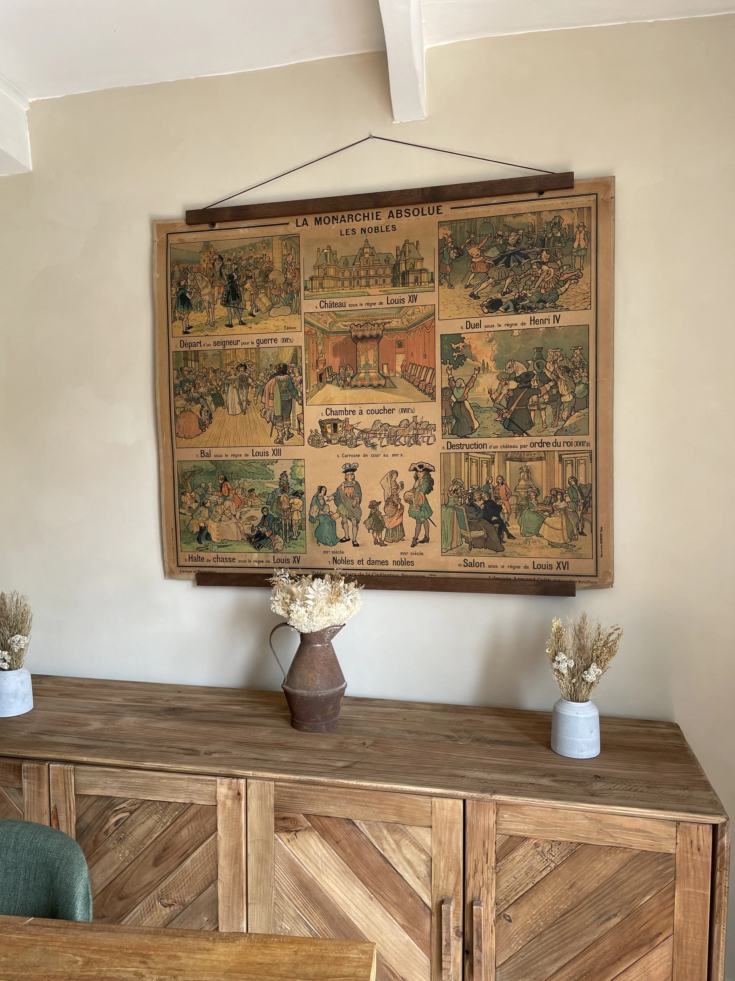 Decorative art print with French text titled "La Monarchie Absolue" hanging above a wooden sideboard. The print features illustrations of French historical scenes and figures from the 16th and 17th centuries. The sideboard has two vases with dried fl