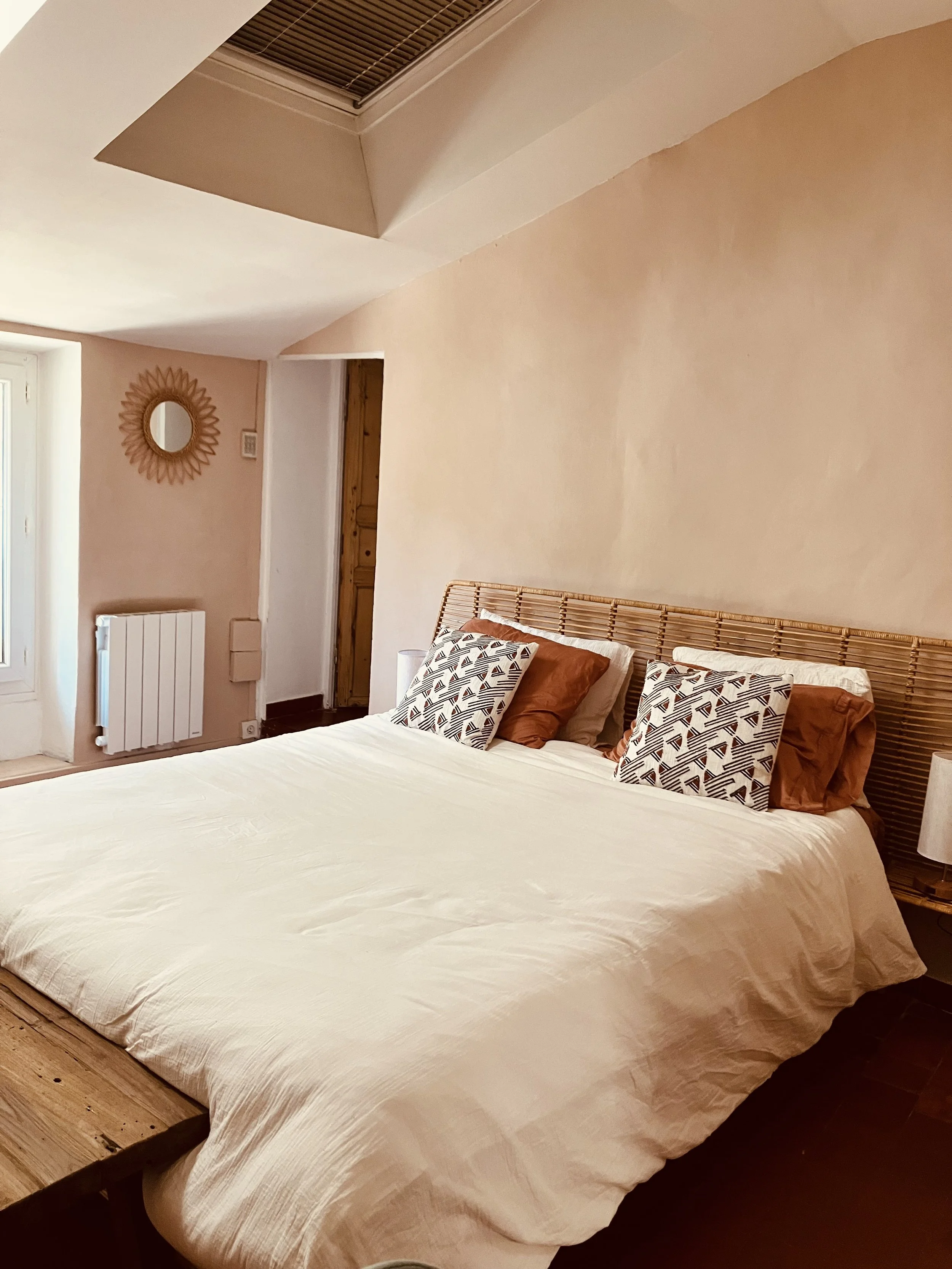 A cozy bedroom with a large bed, beige bedding, patterned pillows, a wooden bed frame, a nightstand with a lamp, and a decorative round mirror on the wall.