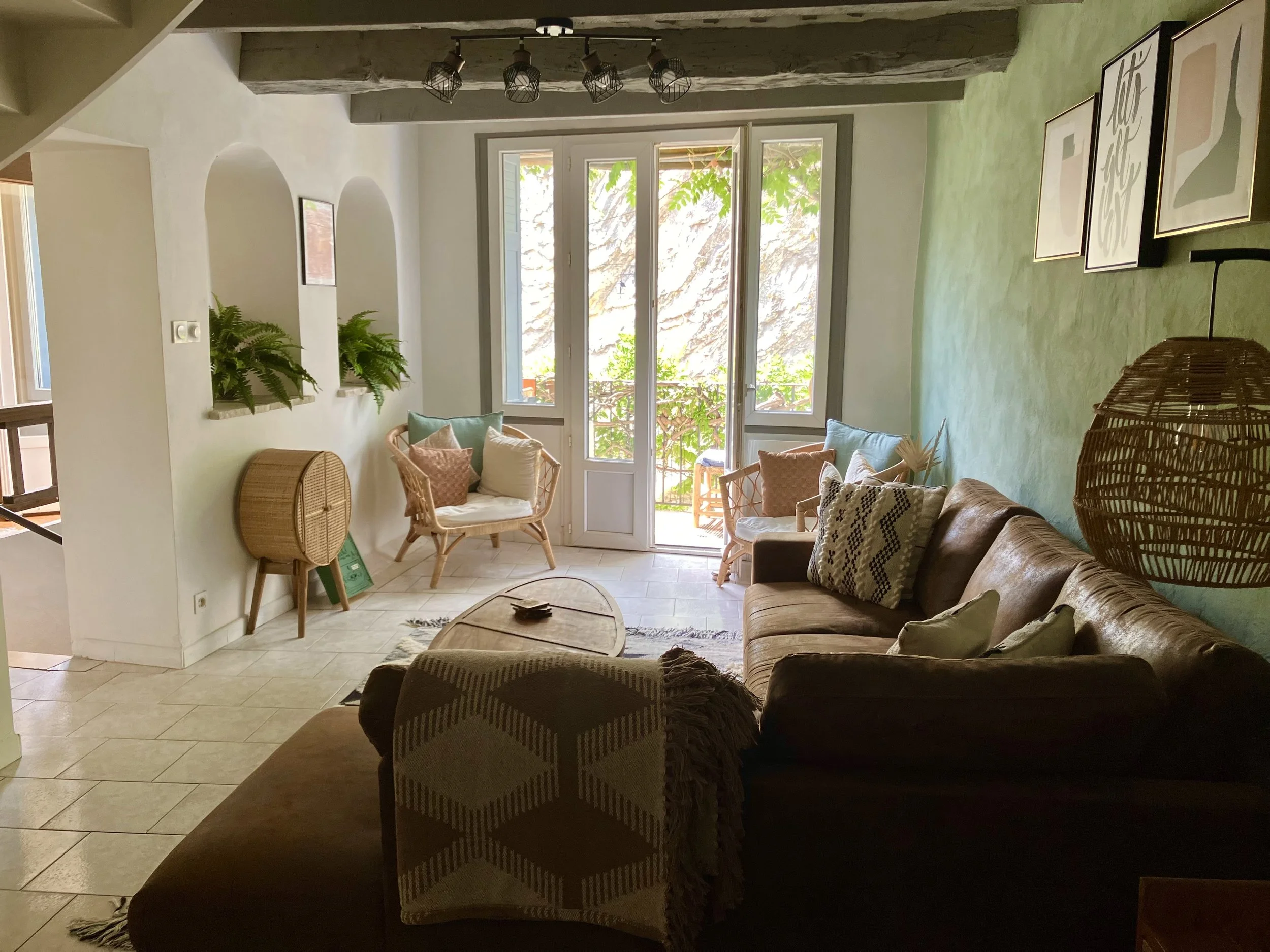 Living room with a green accent wall, wicker chairs with cushions near a large window, framed artwork on the wall, and a beige sofa with patterned pillows.
