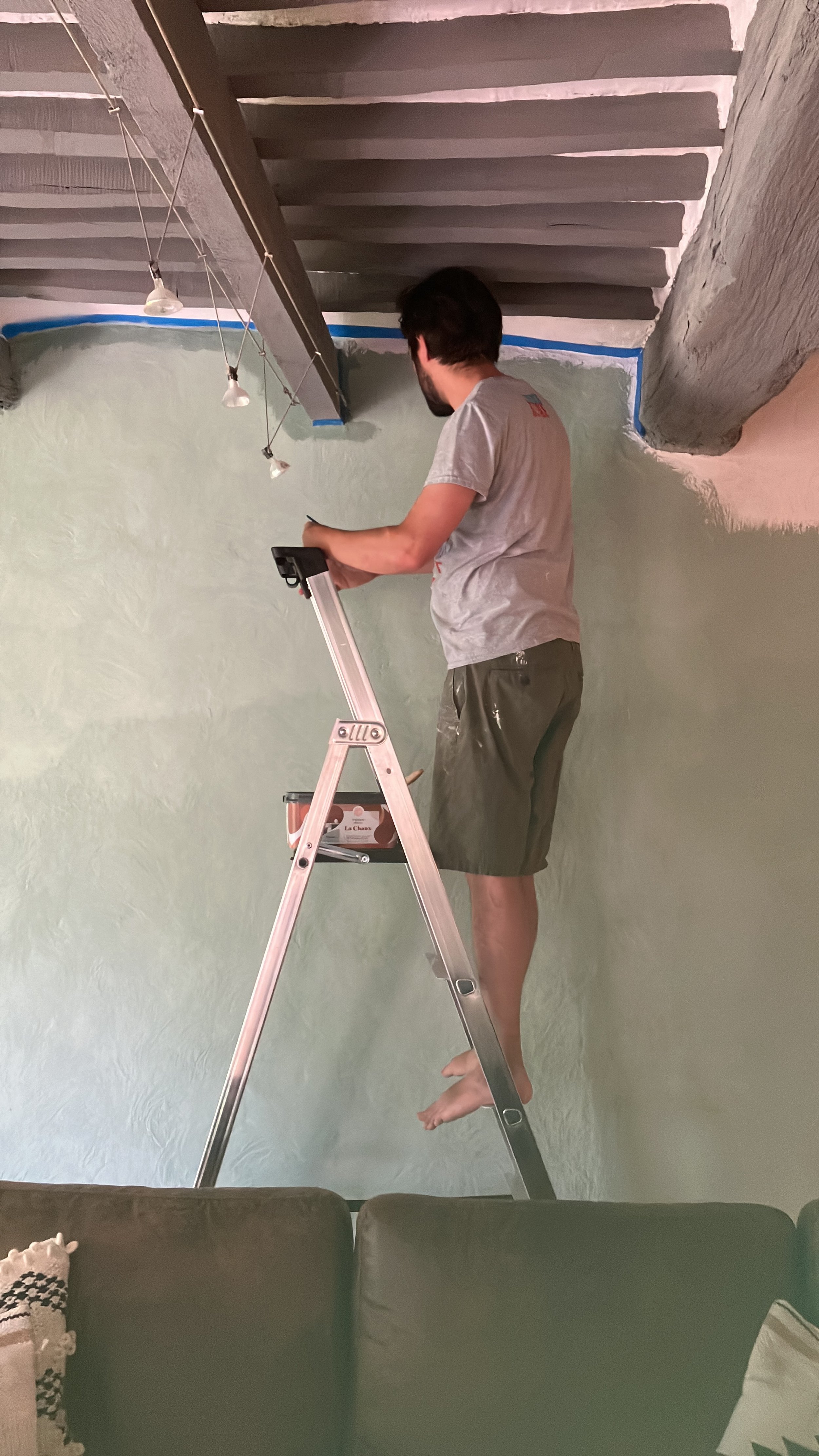A man is standing on a step ladder, painting the upper part of a wall in a room. The room has textured light green walls, and the man is using a paintbrush with paint on it. He is wearing a gray T-shirt and shorts and is barefoot. There is a couch wi