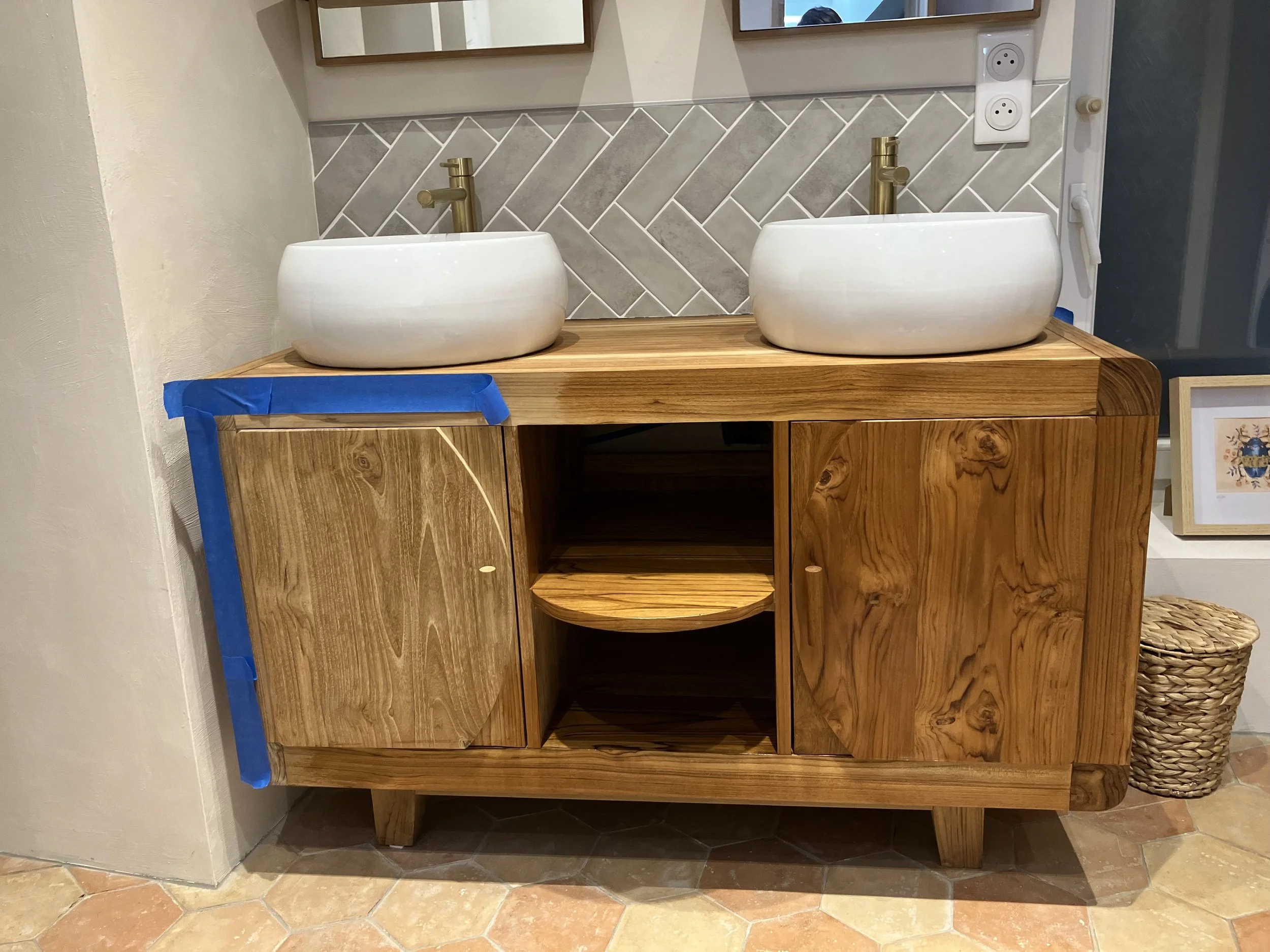 Two white oval vessel sinks with brushed gold faucets sit on a wooden vanity with open storage underneath. The backsplash features a geometric tile pattern. There is a mirror above the sinks and a power outlet to the right.