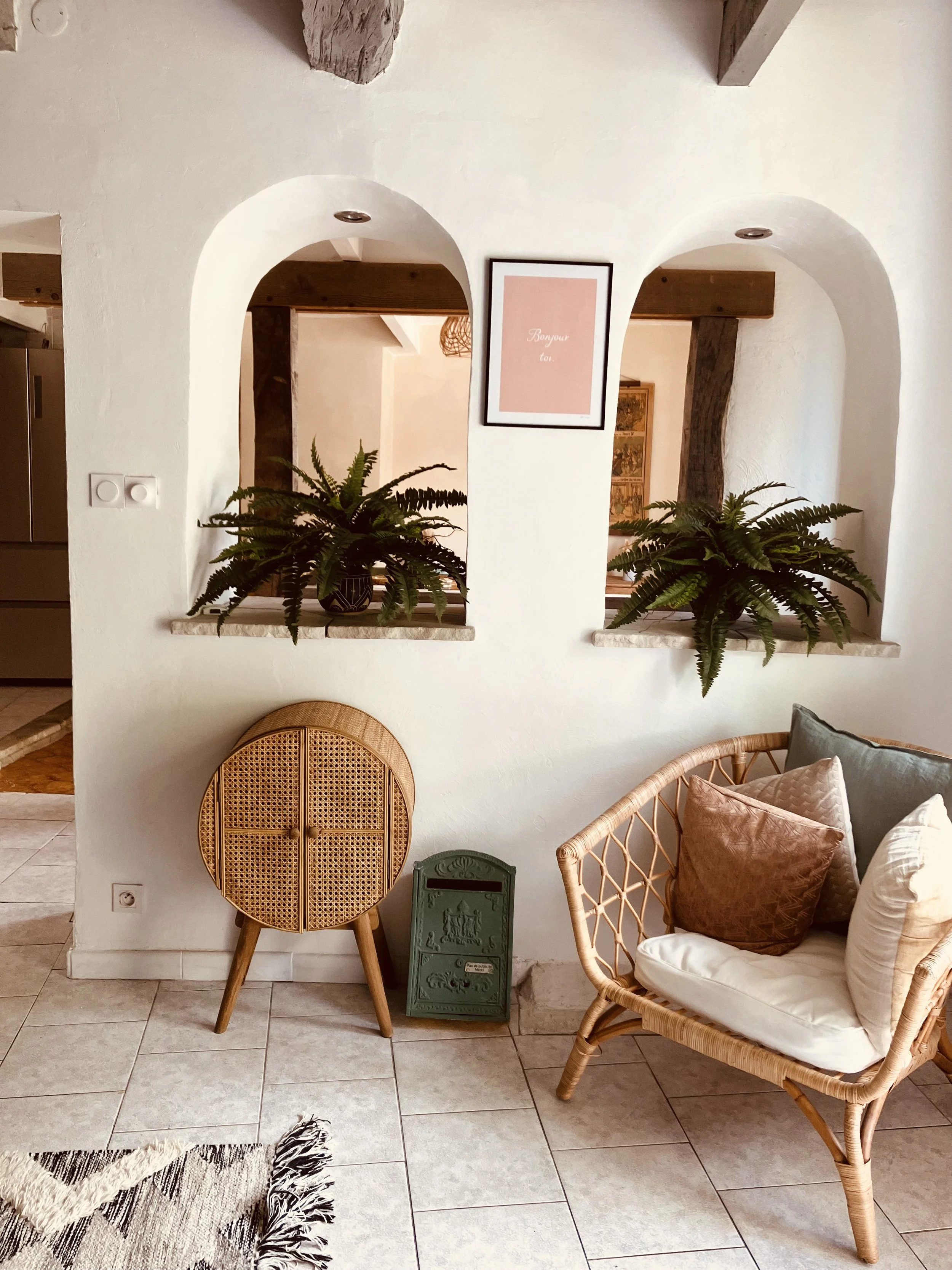 The image shows a cozy living space with a white wall featuring two arched openings. There are two leafy green plants on the window sills, a framed pink print in the center, a rattan chair, and various cushions on a light-colored sofa or chair. There