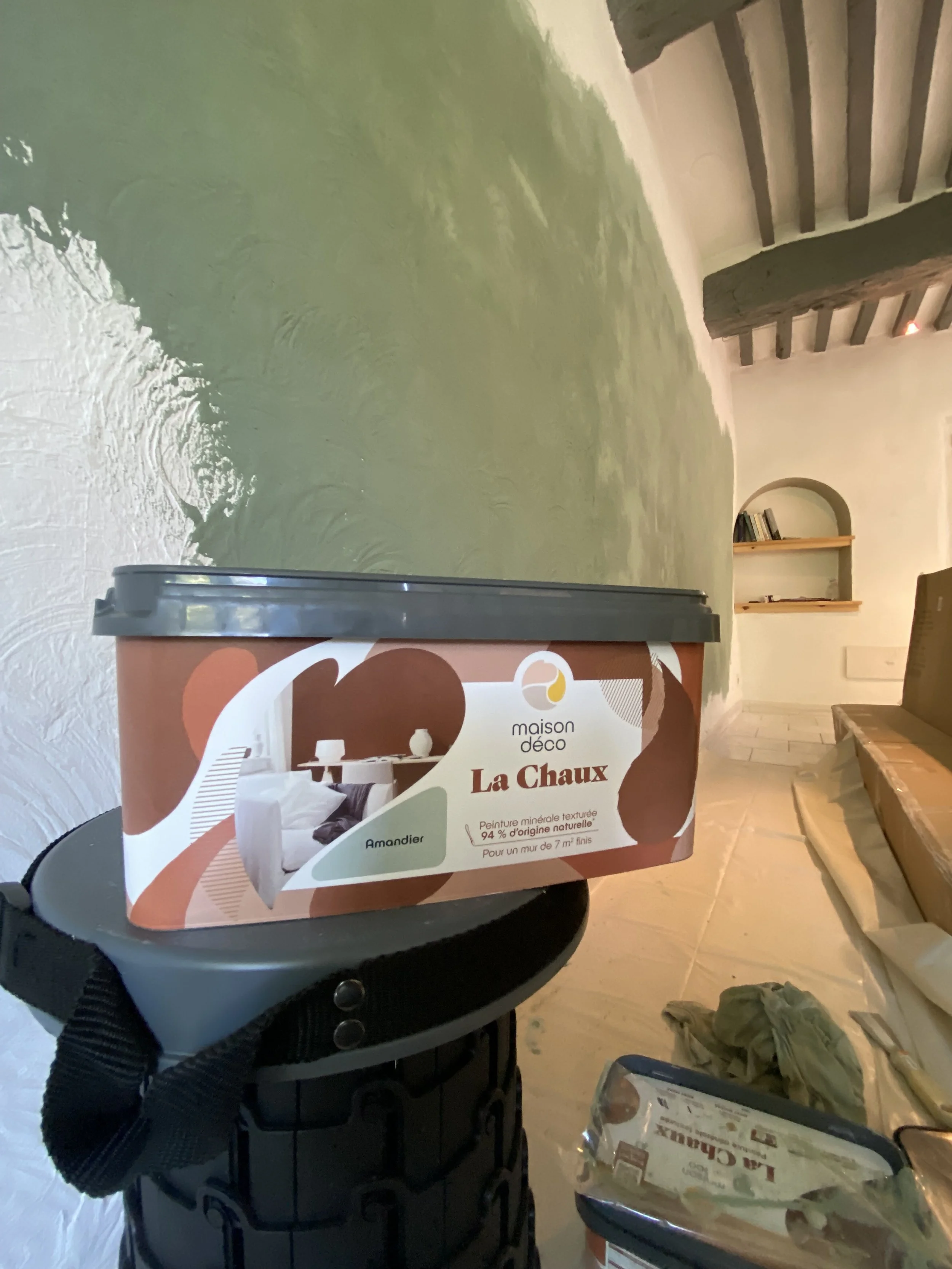 A person holding a box of latex paint labeled 'La Chaux' in a room under renovation. The room has wooden stairs, a beige bench, and scattered construction supplies.
