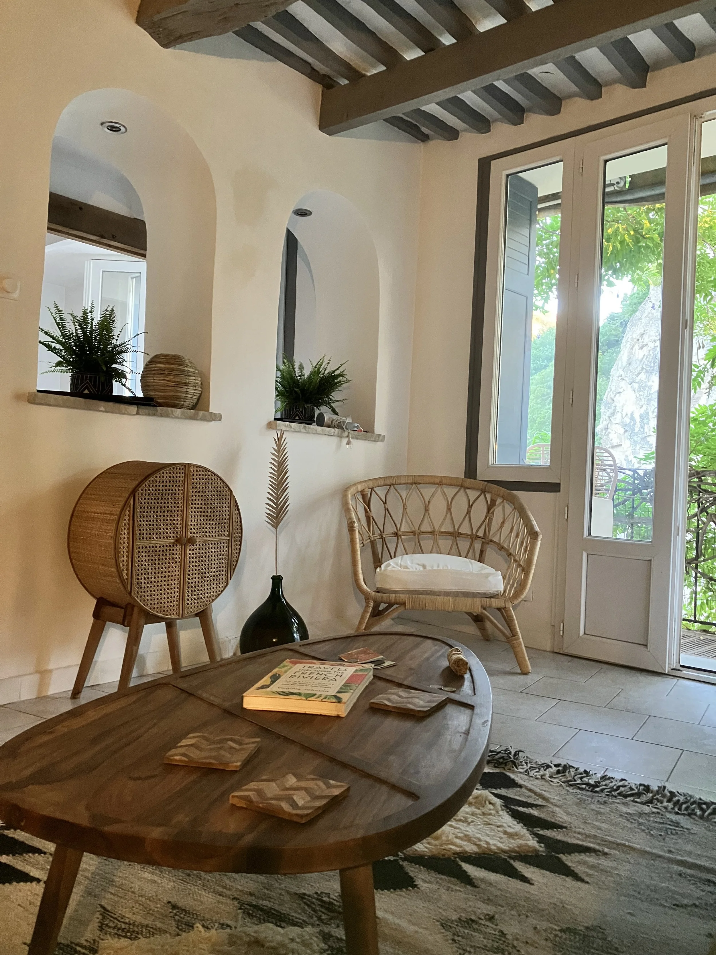 Living room with a large glass door opening to a balcony showing trees outside. The room features woven chairs, a round wooden coffee table with magazines and coasters, potted plants in black and woven pots, a rug, and some decorative items.