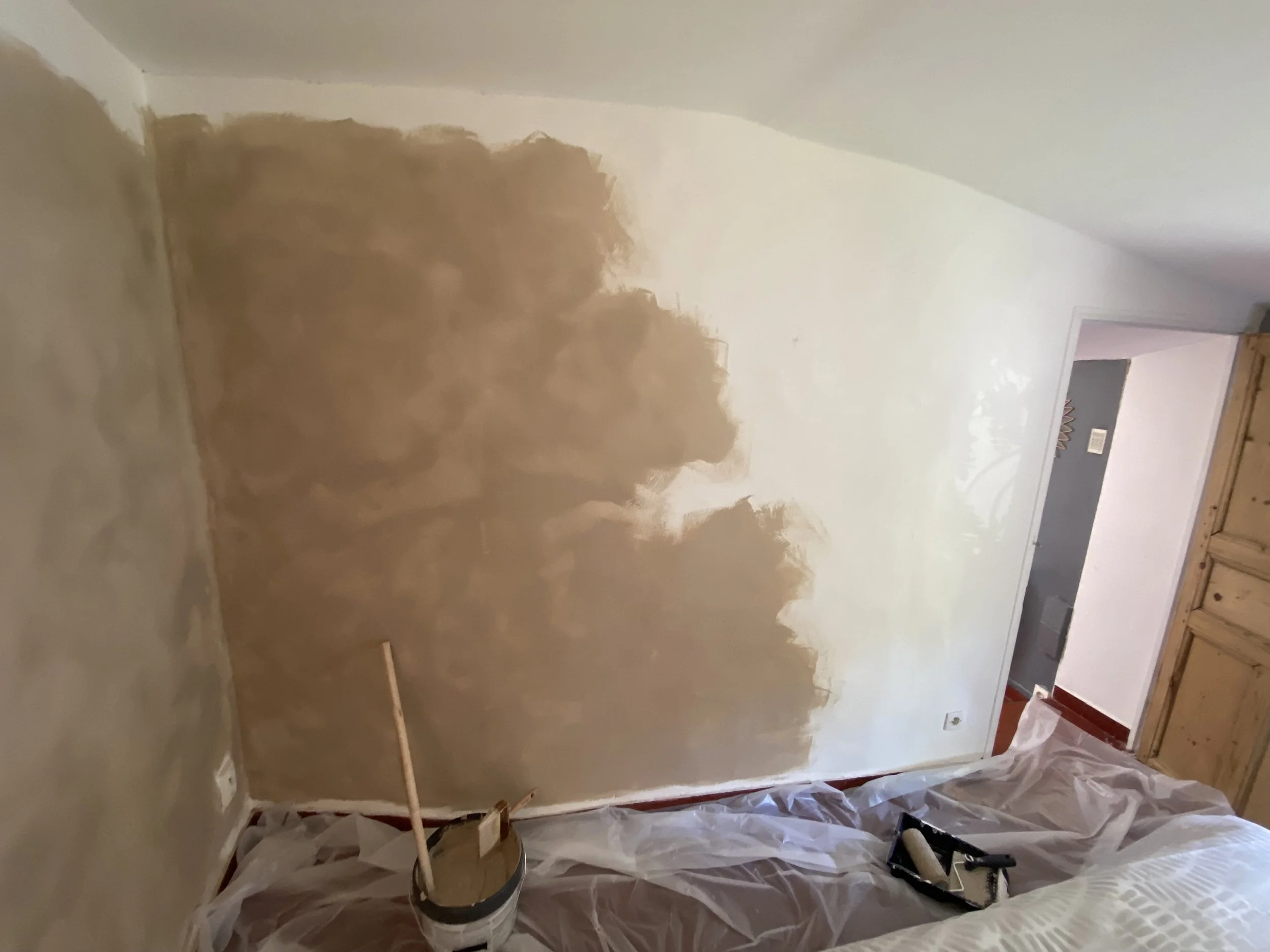 Room with partially painted walls, painting supplies, and plastic sheeting on the floor.