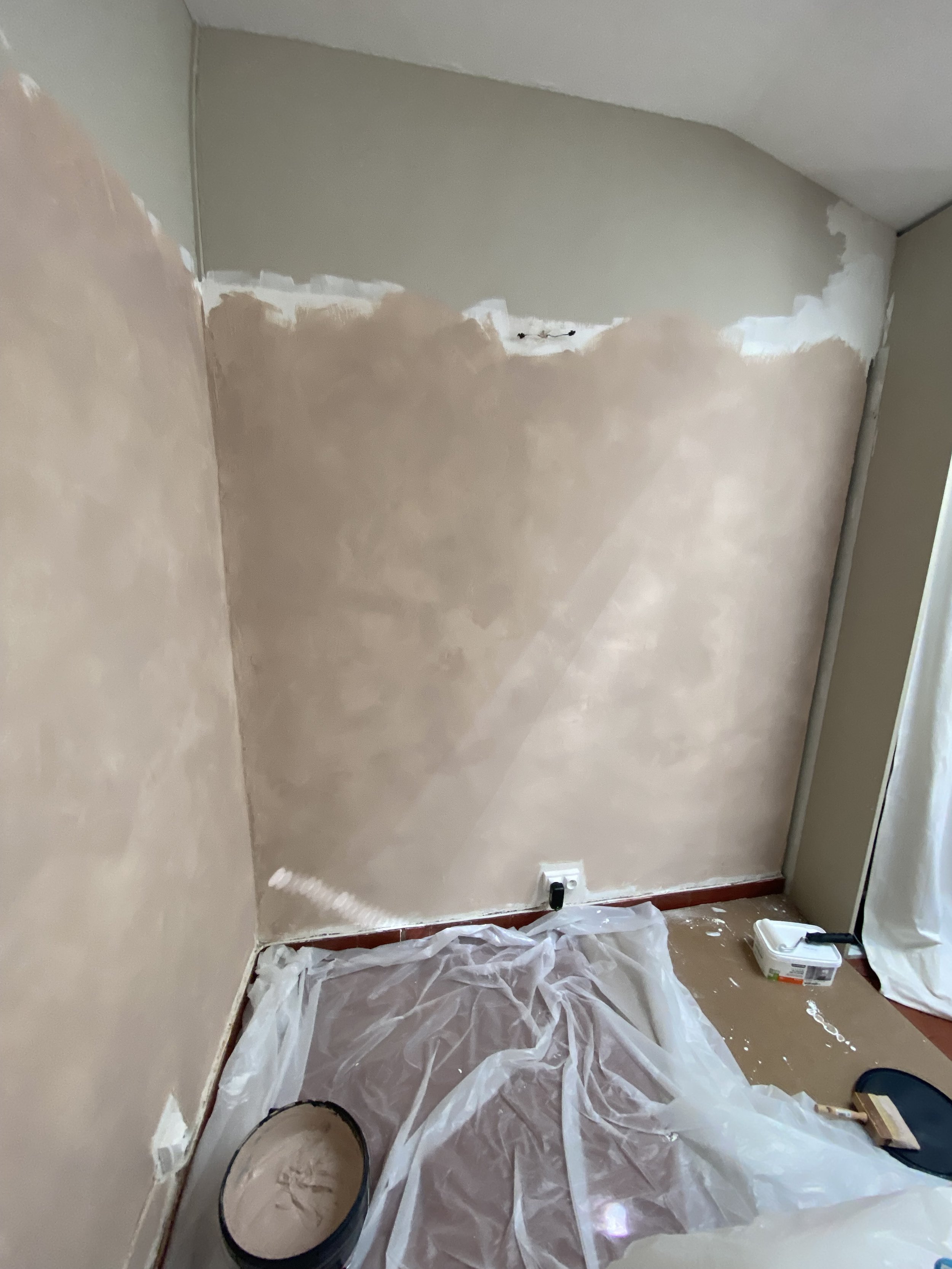 An interior wall being painted, with plastic sheeting covering the floor, a bucket of paint, and painting supplies nearby.