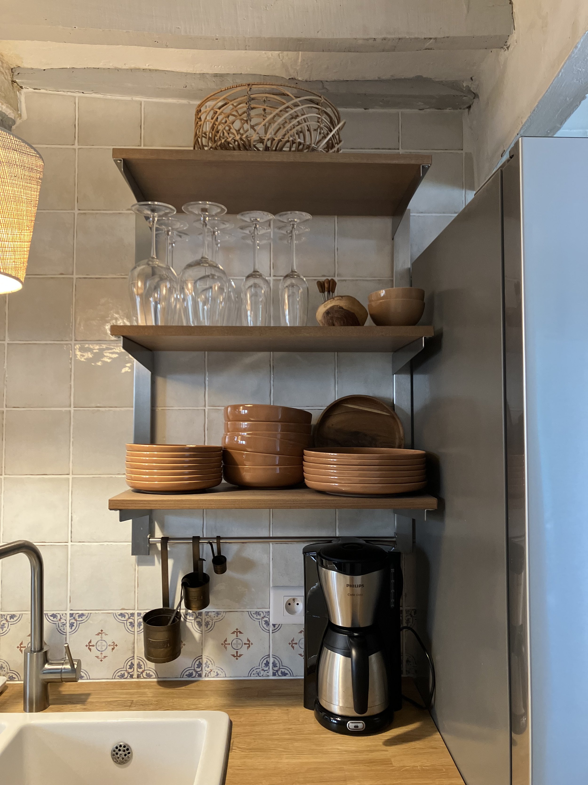 A kitchen counter with a wooden surface, a coffee grinder, and a shelf with glasses, bowls, and mugs. The wall behind is tiled with beige and blue patterns, and a part of a sink is visible in the foreground.