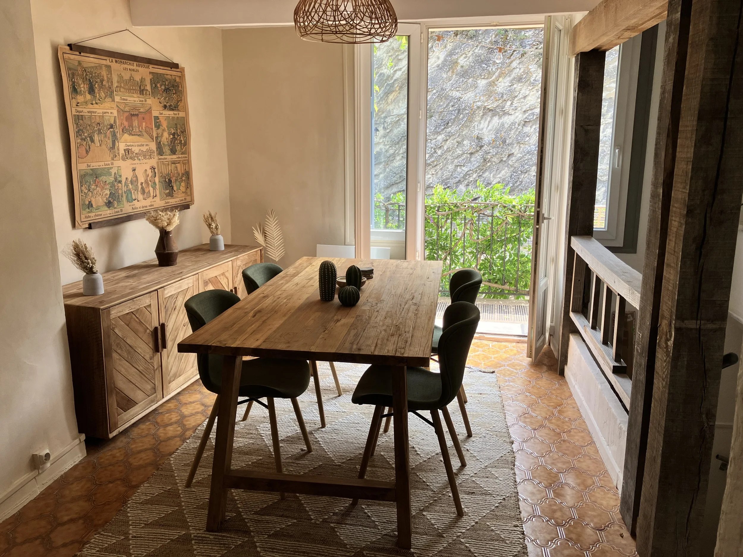 A dining room with a wooden table and six black chairs, a wooden sideboard with vases, a large picture on the wall, and a glass door leading to a balcony with green foliage outside.
