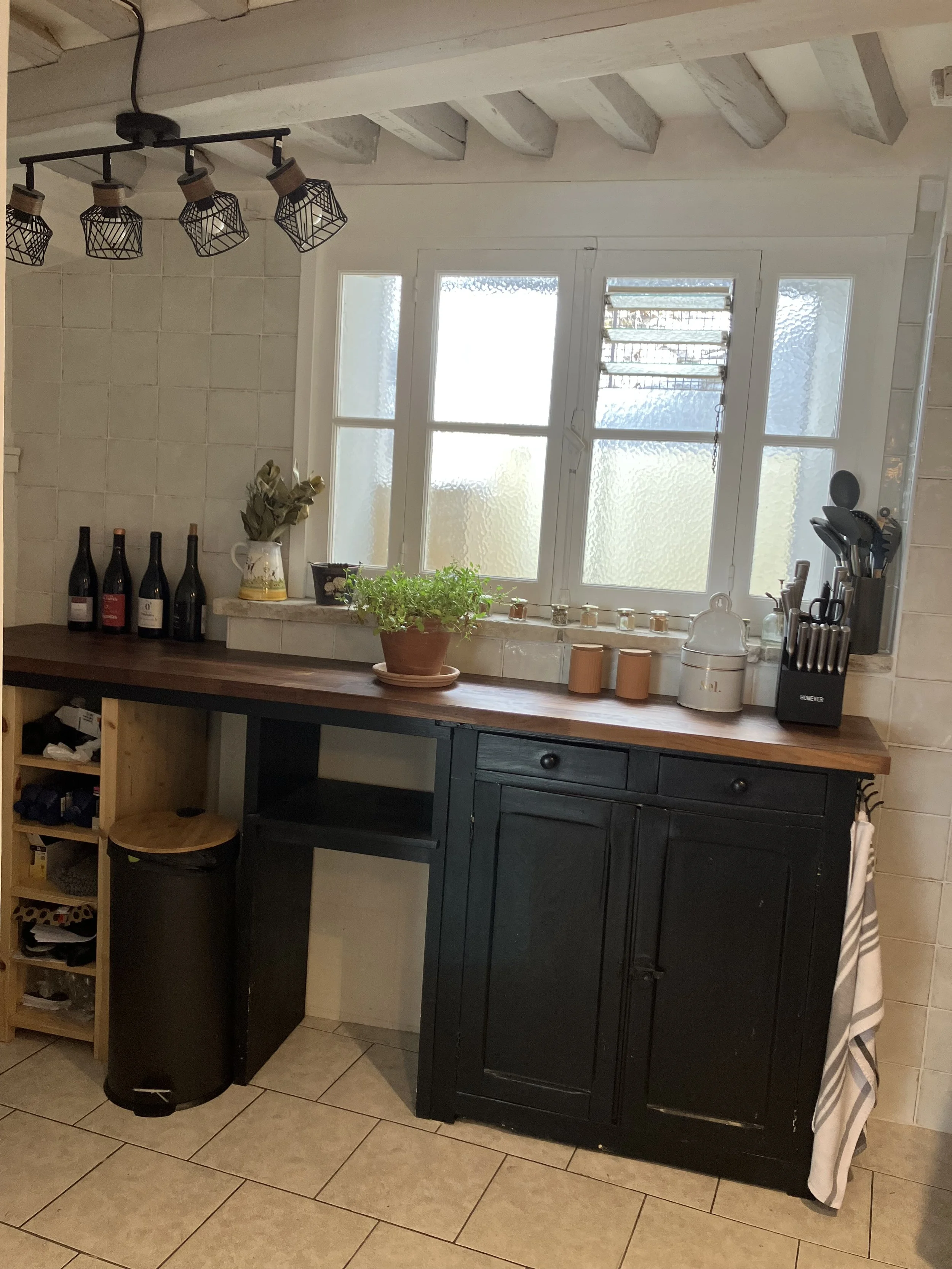 A kitchen window with frosted glass, a black countertop with bottles, potted plants, jars, kitchen utensils, and a black cabinet, with a small wooden shelf, a trash bin, and a towel on a hook nearby.