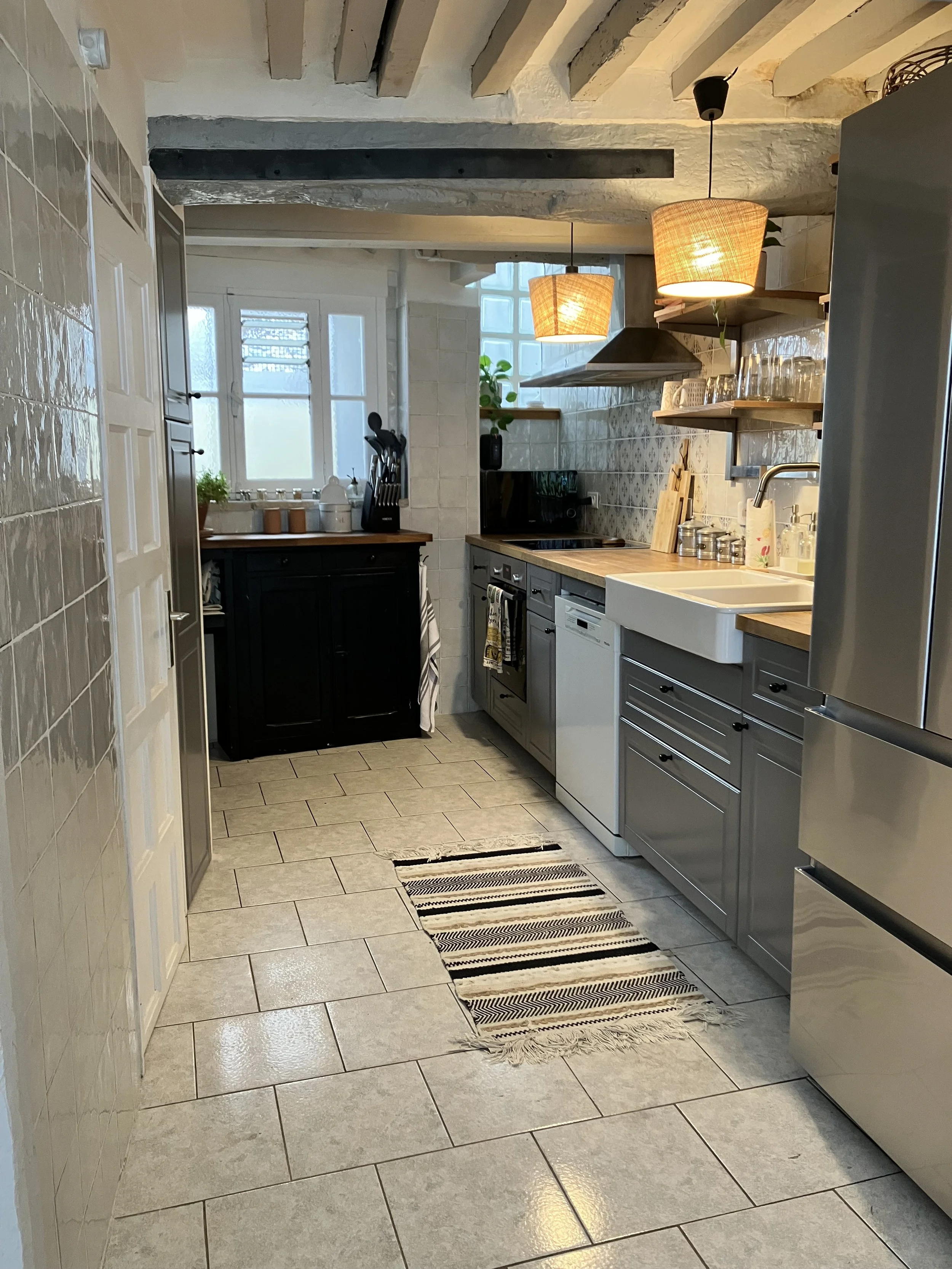 A cozy kitchen with beige tiled floor, a gray cabinet with black drawers, a white sink, stainless steel appliances, open wooden shelves holding glassware, a black microwave, a wooden cutting board, and two hanging wicker lamps. There is a window with